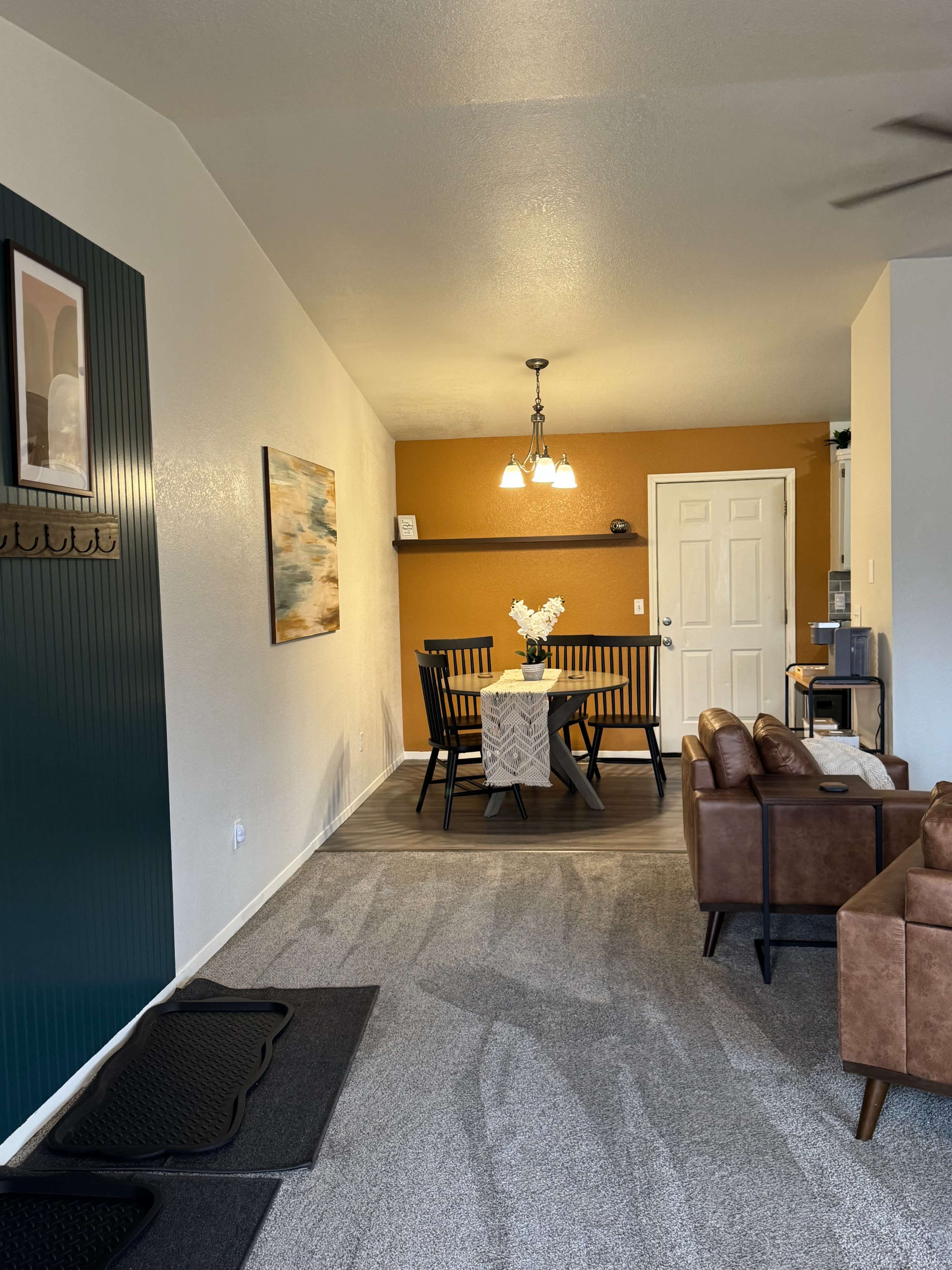 The image shows a living space with a carpeted floor leading to a dining area, featuring a table with chairs and an orange accent wall.