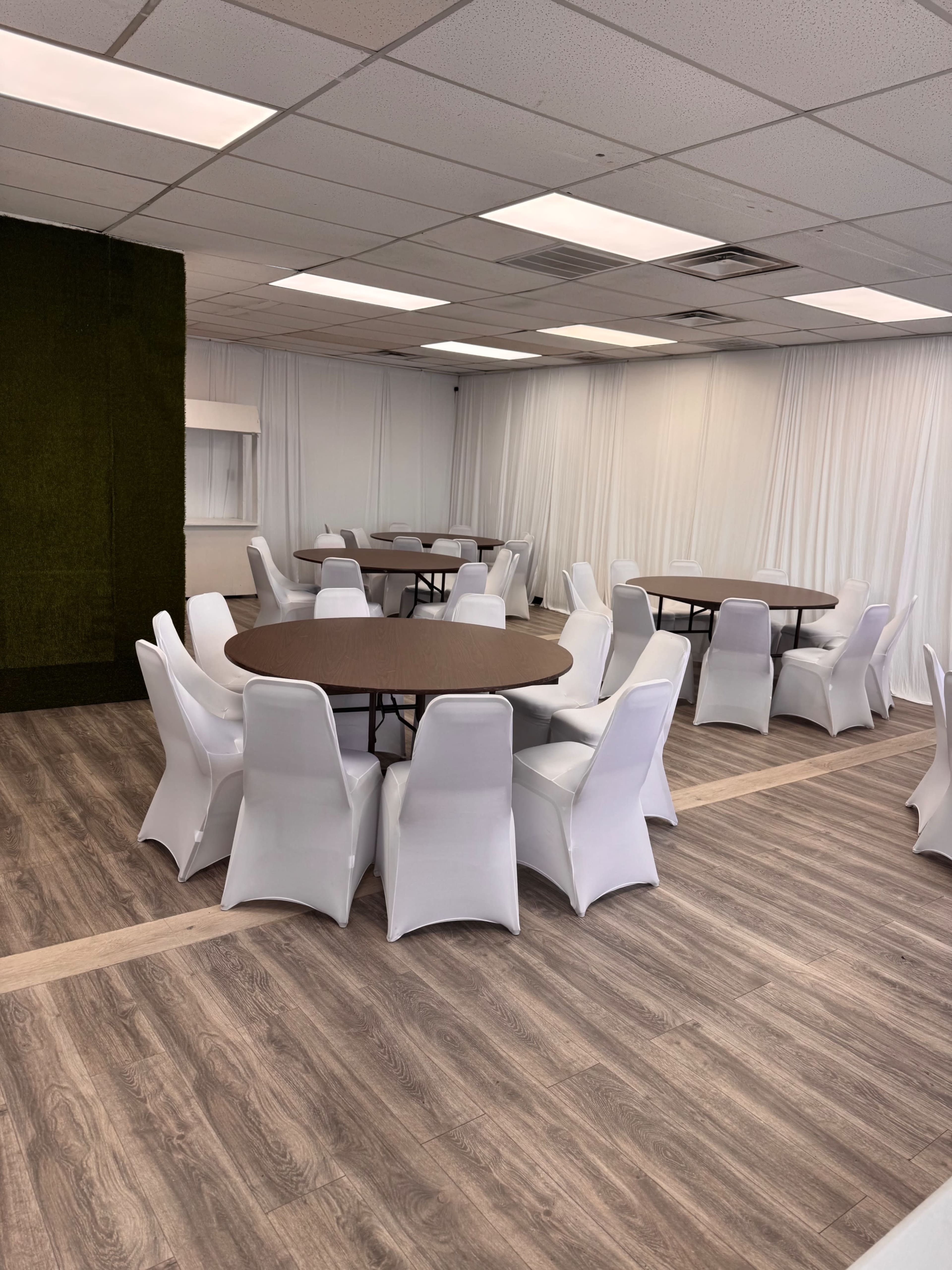 The image shows a spacious event room with several round tables surrounded by white folding chairs and beige wood-like flooring.