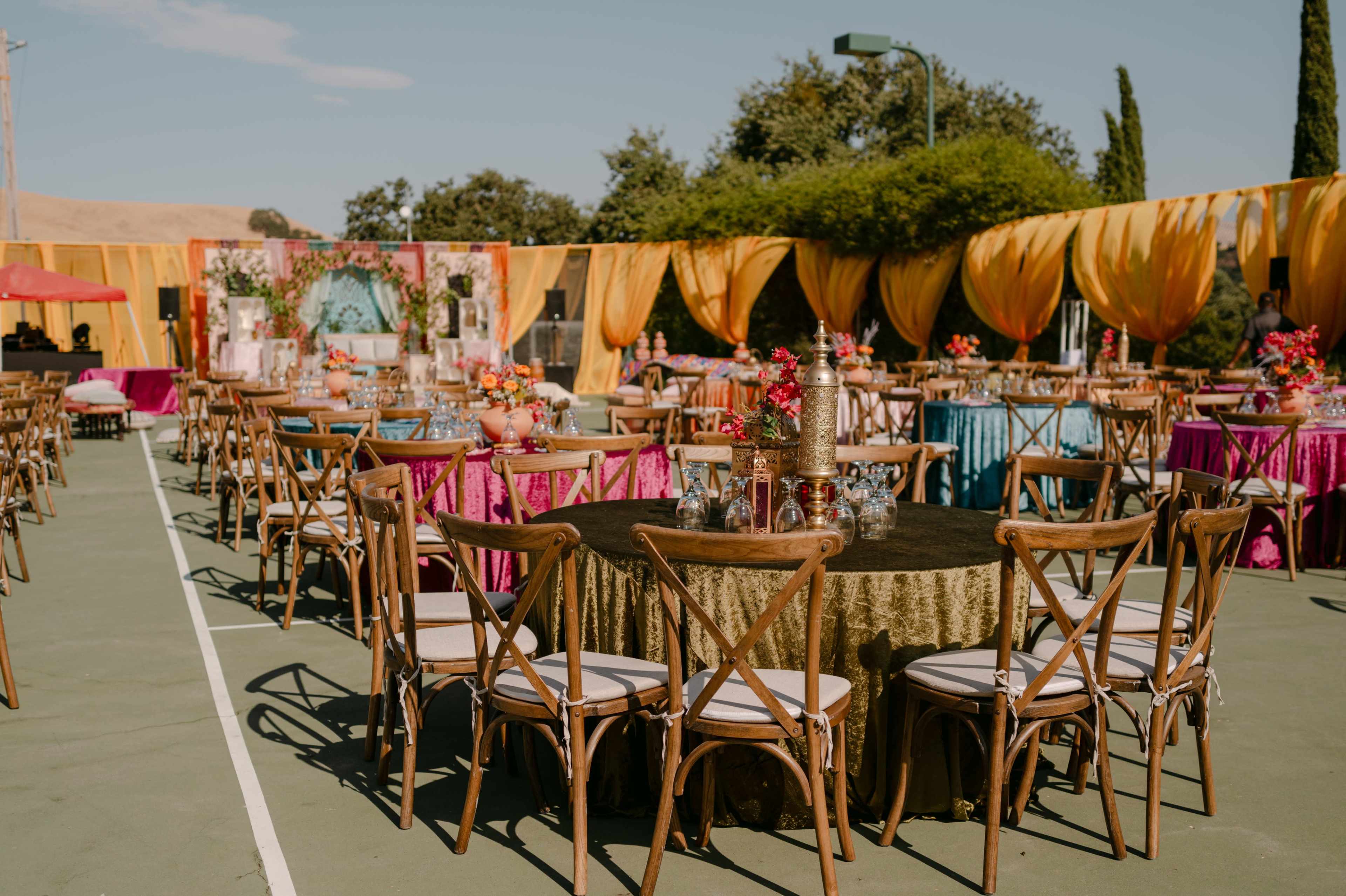 The image shows a decorated outdoor event space with multiple tables covered in colorful tablecloths, surrounded by chairs, and adorned with floral arrangements.