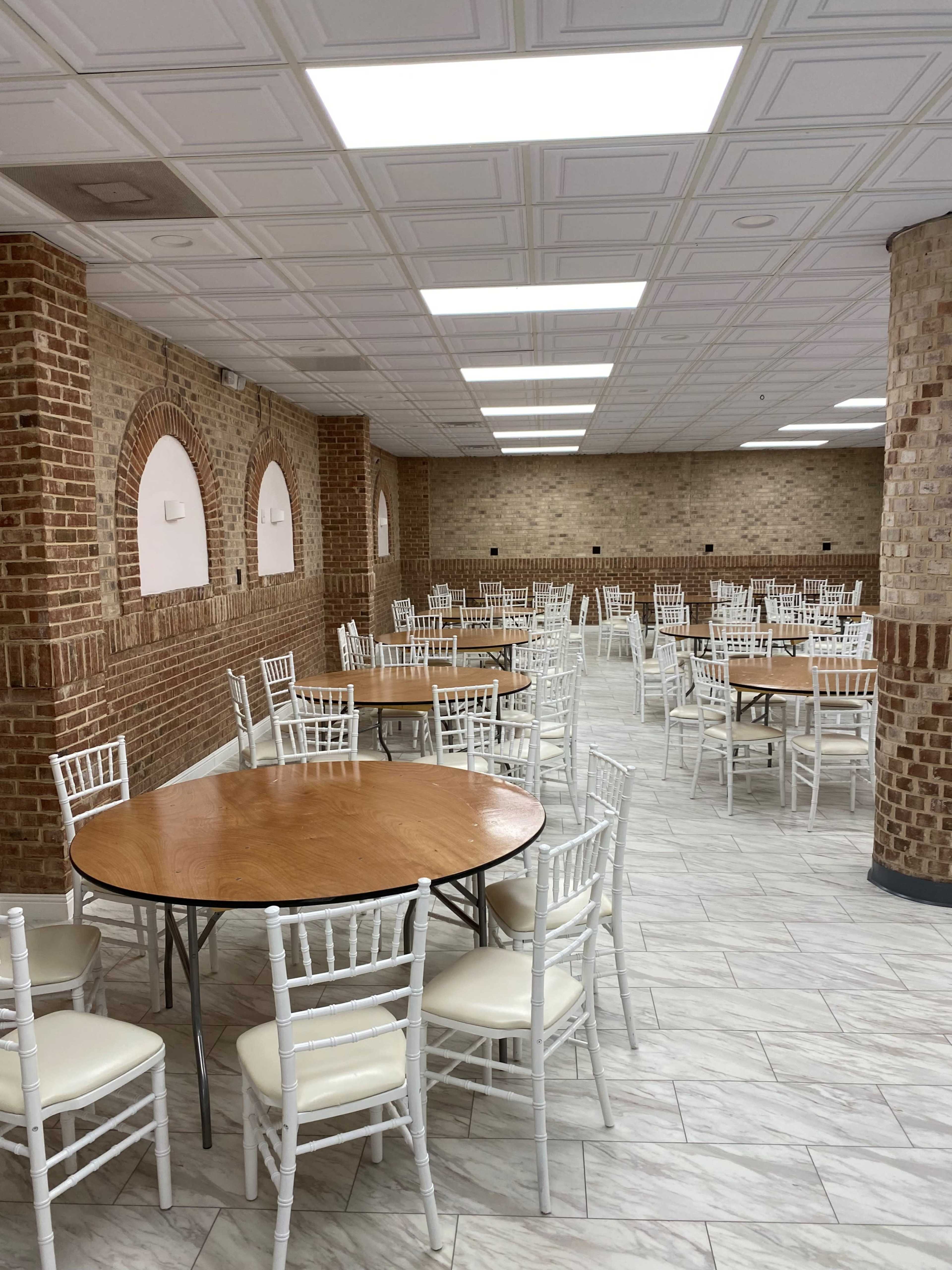 A large, empty dining area features multiple round tables surrounded by white chairs, with brick walls and a tiled floor.