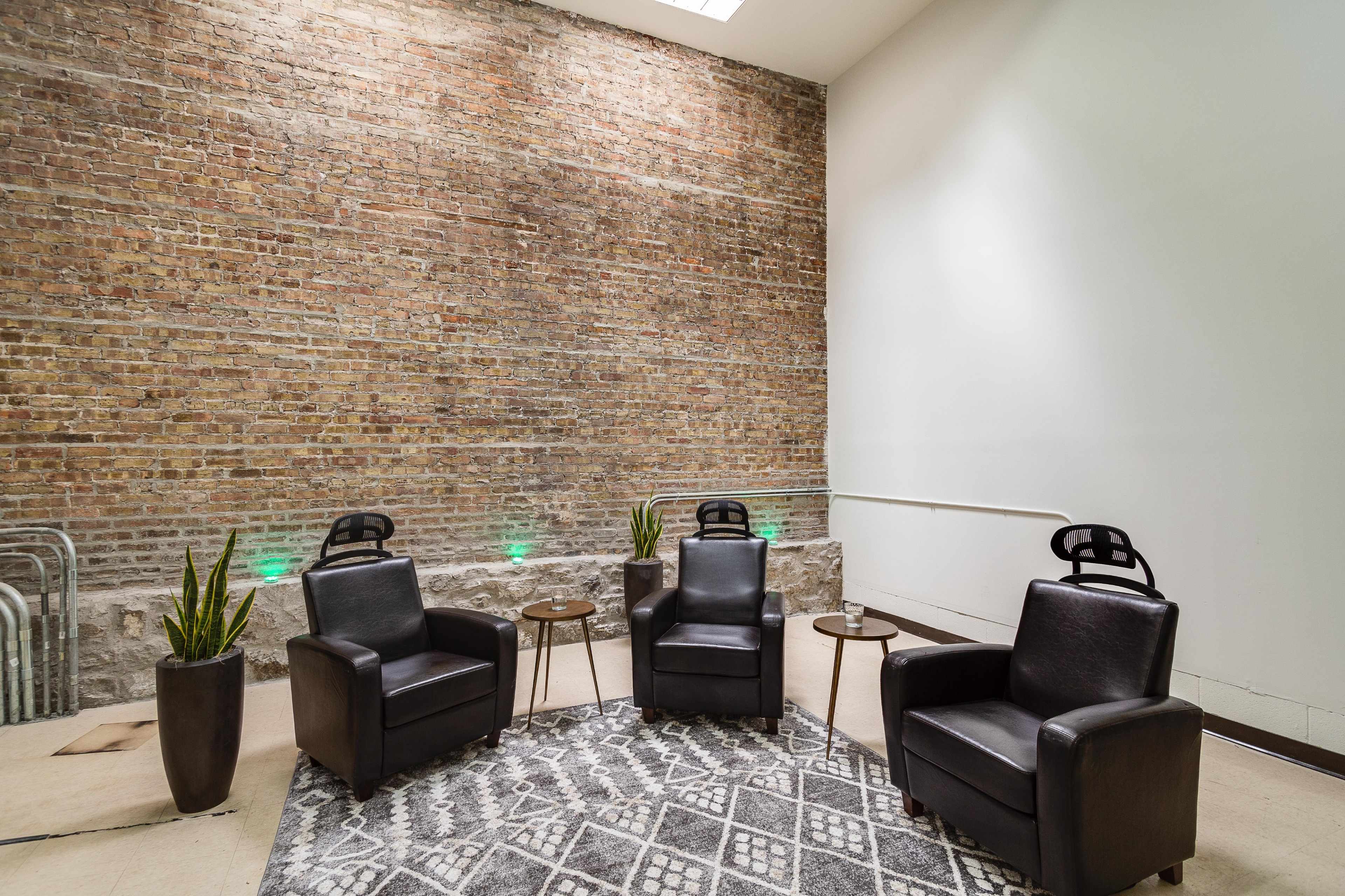A minimalist seating area with two black chairs, small wooden tables, and greenery against a brick wall.