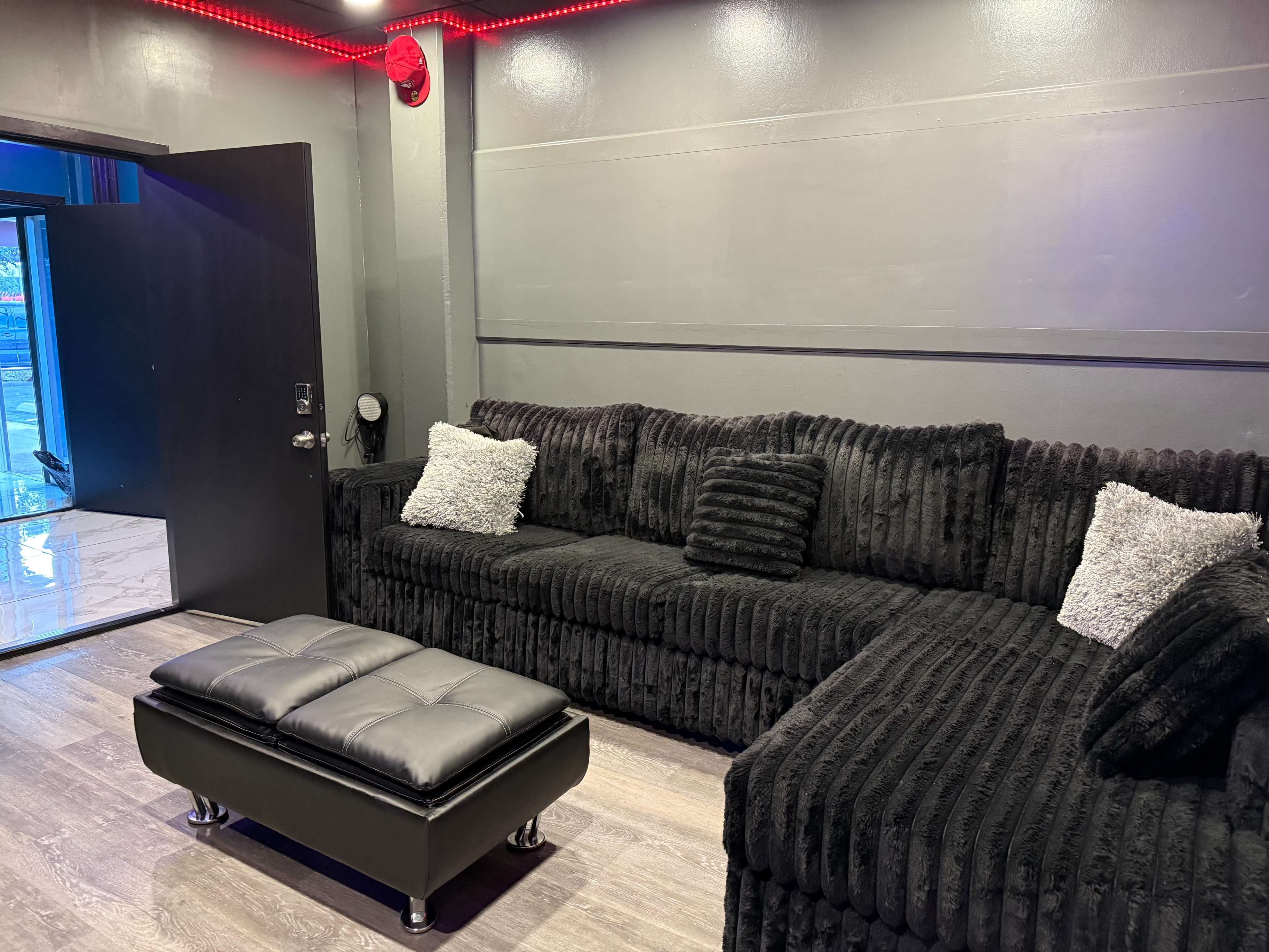 The image shows a modern lounge area with a large black sectional sofa and a matching ottoman in a room with gray walls and ambient lighting.