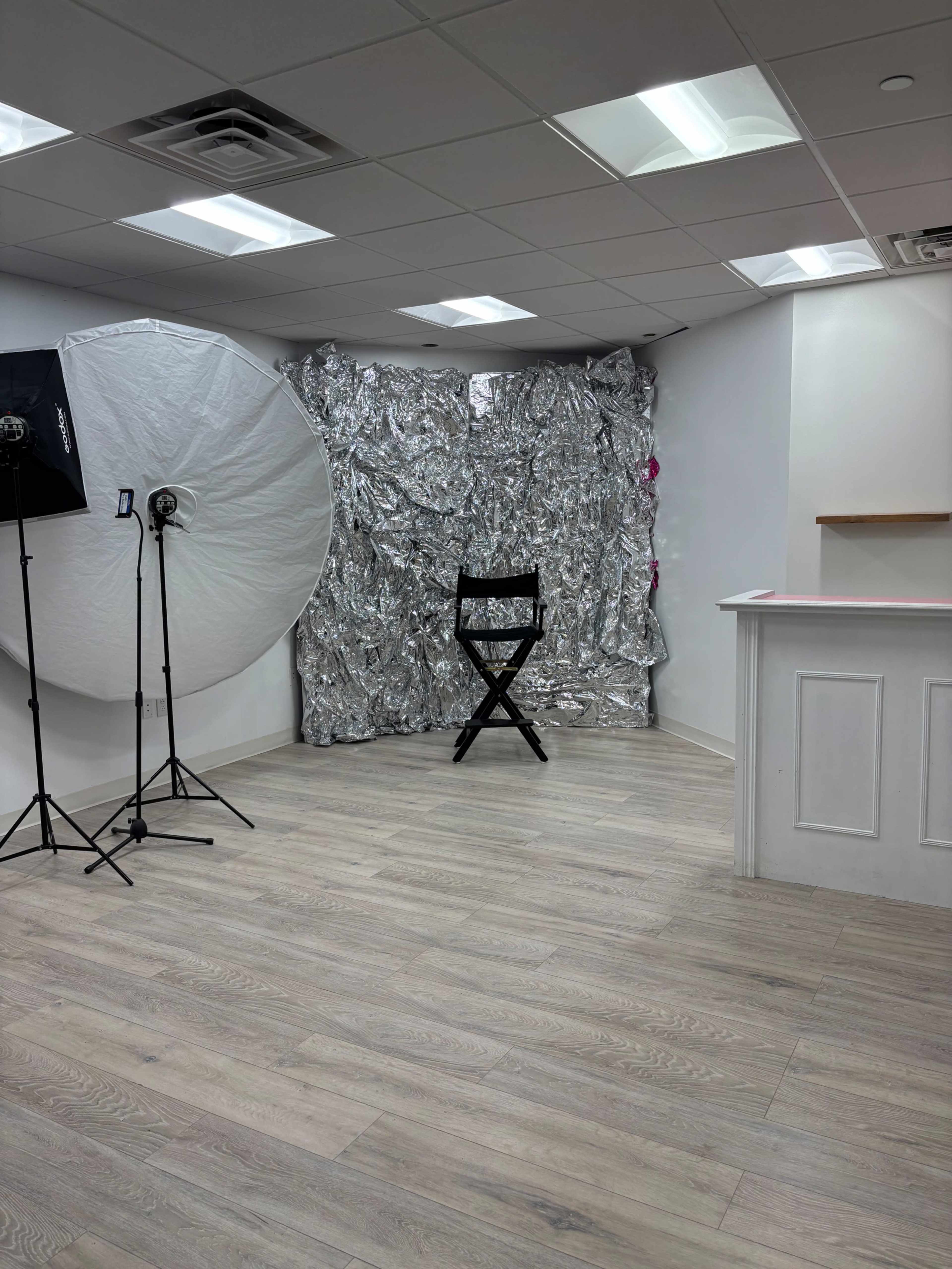 The image shows a photography studio with a silver backdrop, a black director's chair in the center, and lighting equipment in the corner.