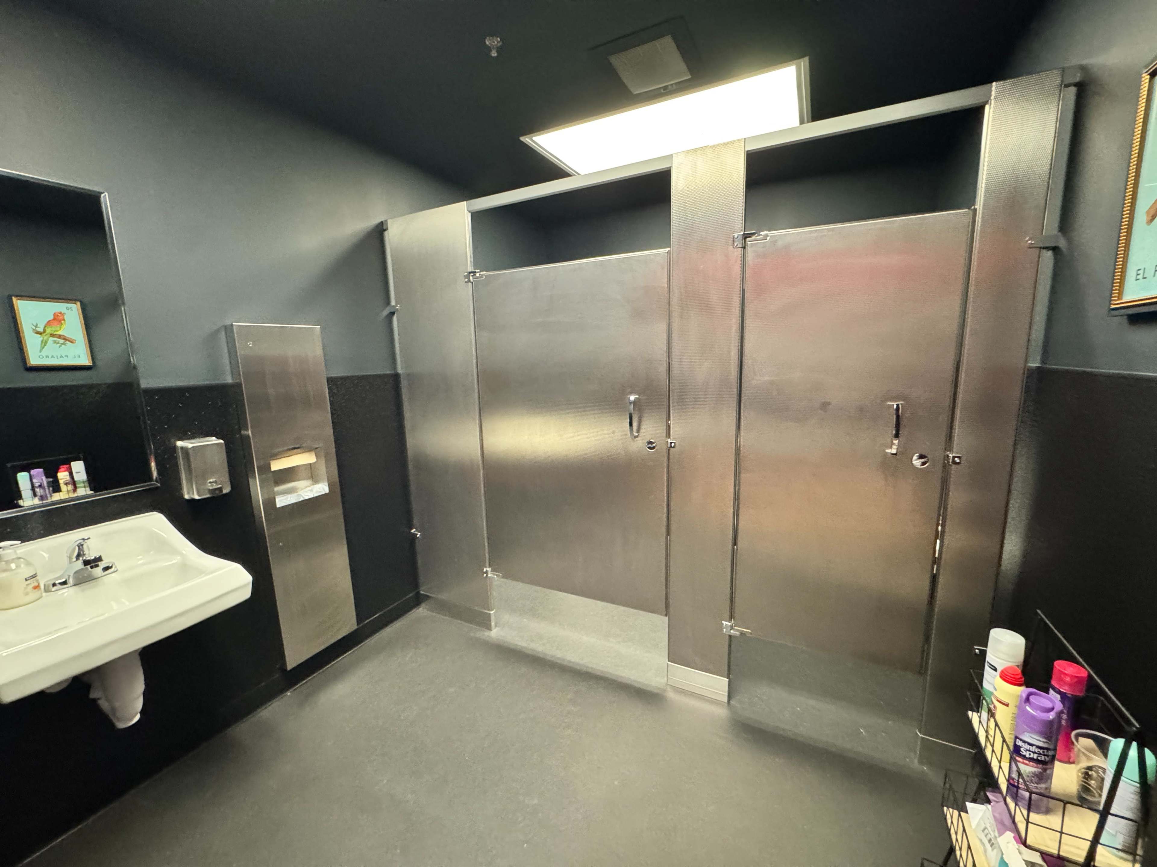 The image shows a clean, dimly lit restroom with metal stall doors, a sink on the left, and various toiletries displayed on a rack.