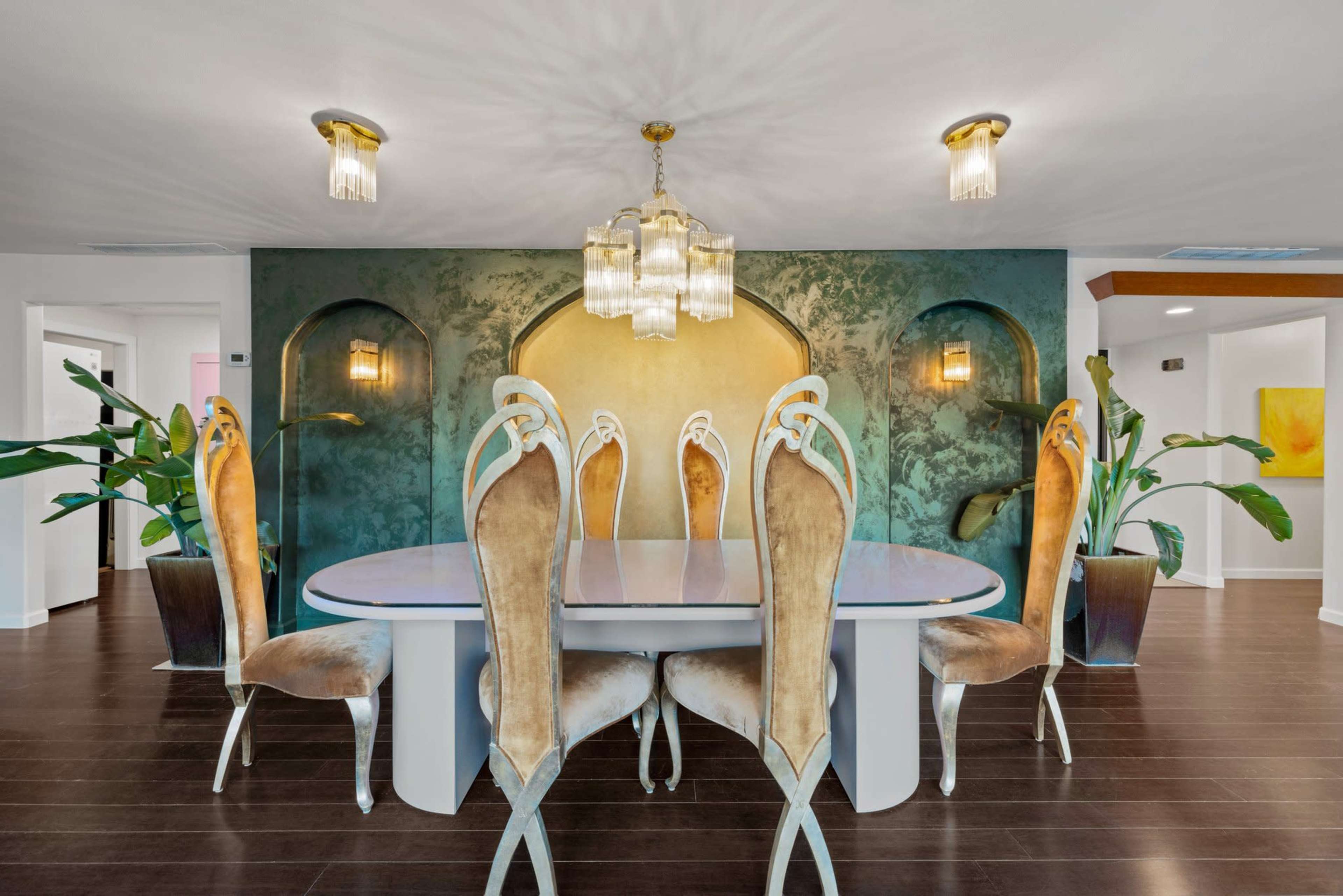 A modern dining room with an oval table surrounded by elegant chairs, set against a textured green and gold wall and accented by indoor plants.