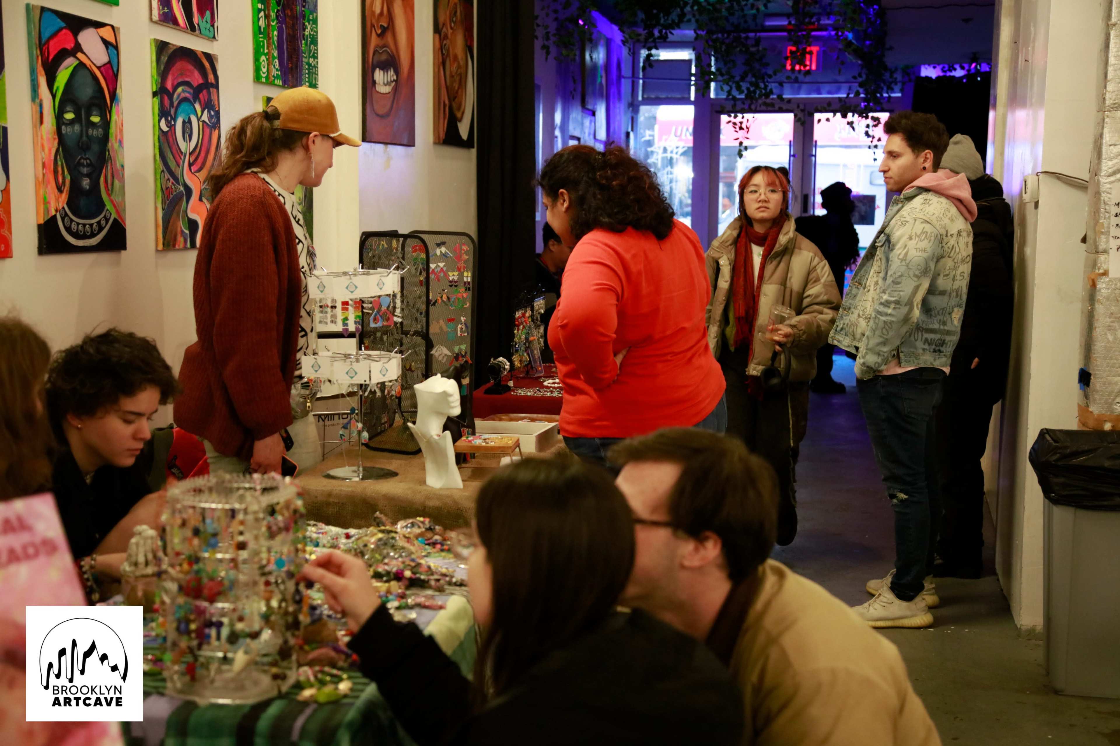 A bustling art gallery setup with vendors displaying handmade jewelry and artworks, while visitors engage in conversation and browse the displays.