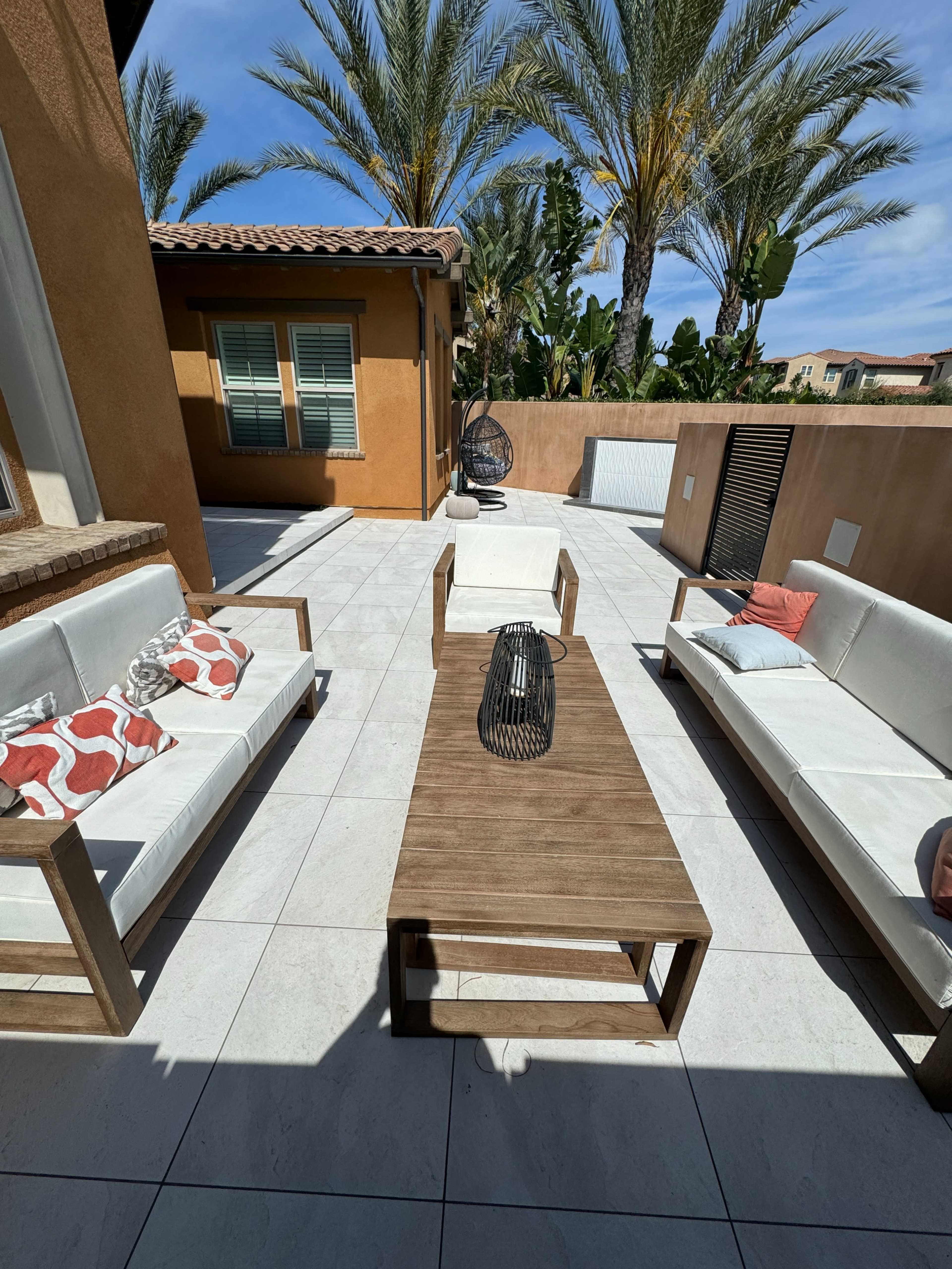 A patio area features two white sofas, a wooden coffee table, and a decorative lantern, surrounded by palm trees and a beige stucco building.