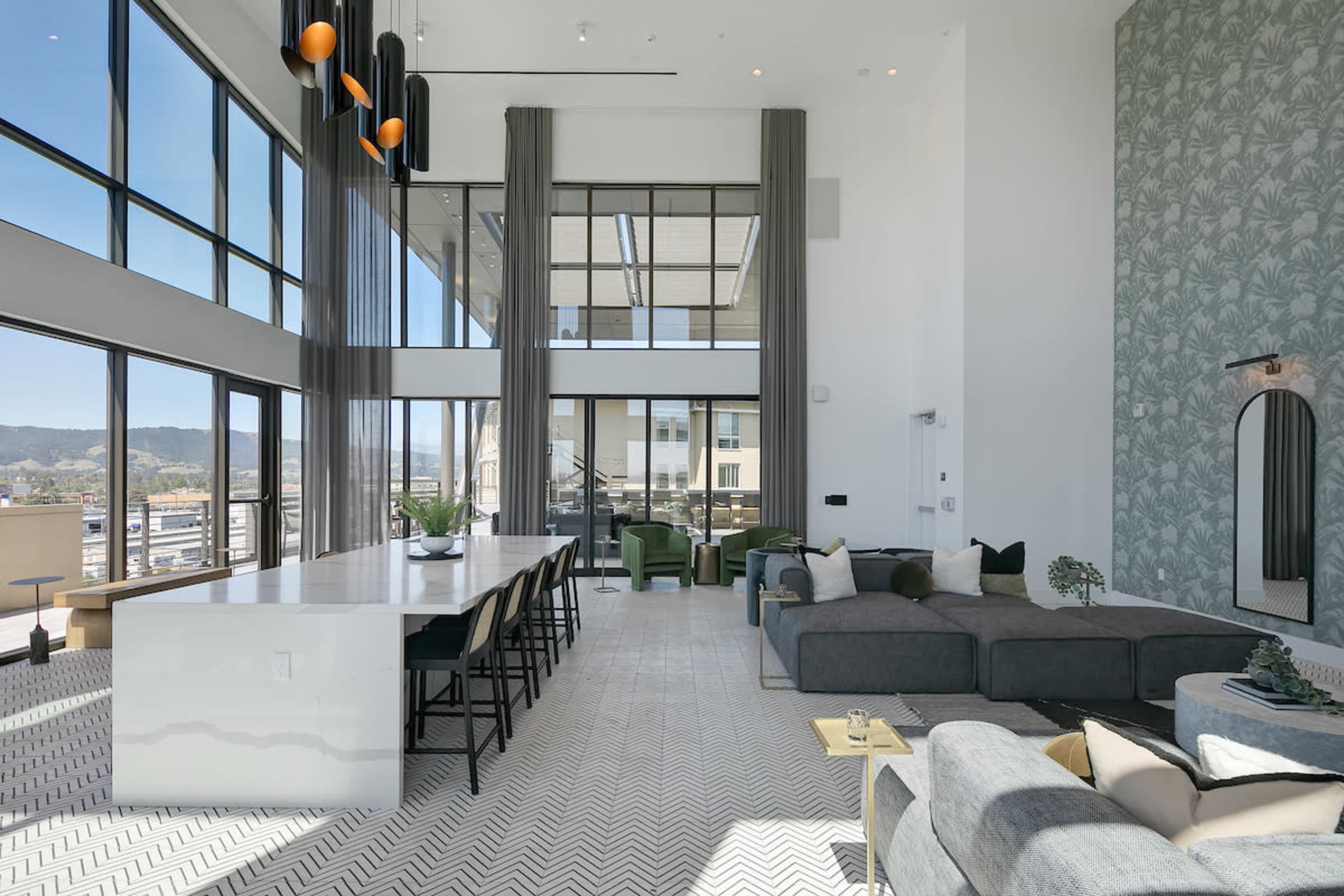 The image shows a spacious, modern living area with large windows, featuring a sectional sofa, a dining table, and decorative plants.