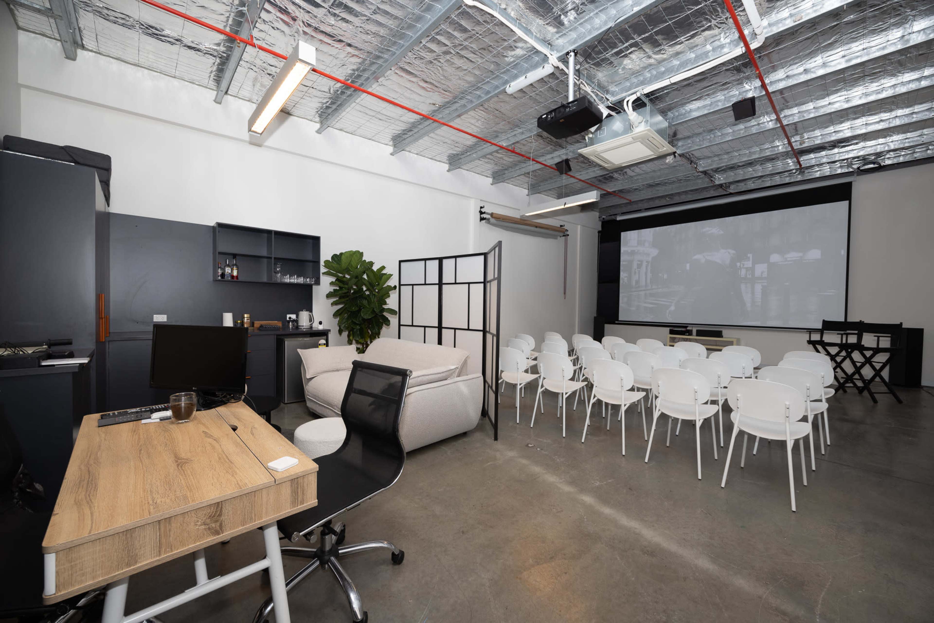 The image shows a modern meeting room with a desk and chair on one side and rows of white chairs facing a large screen on the other side.