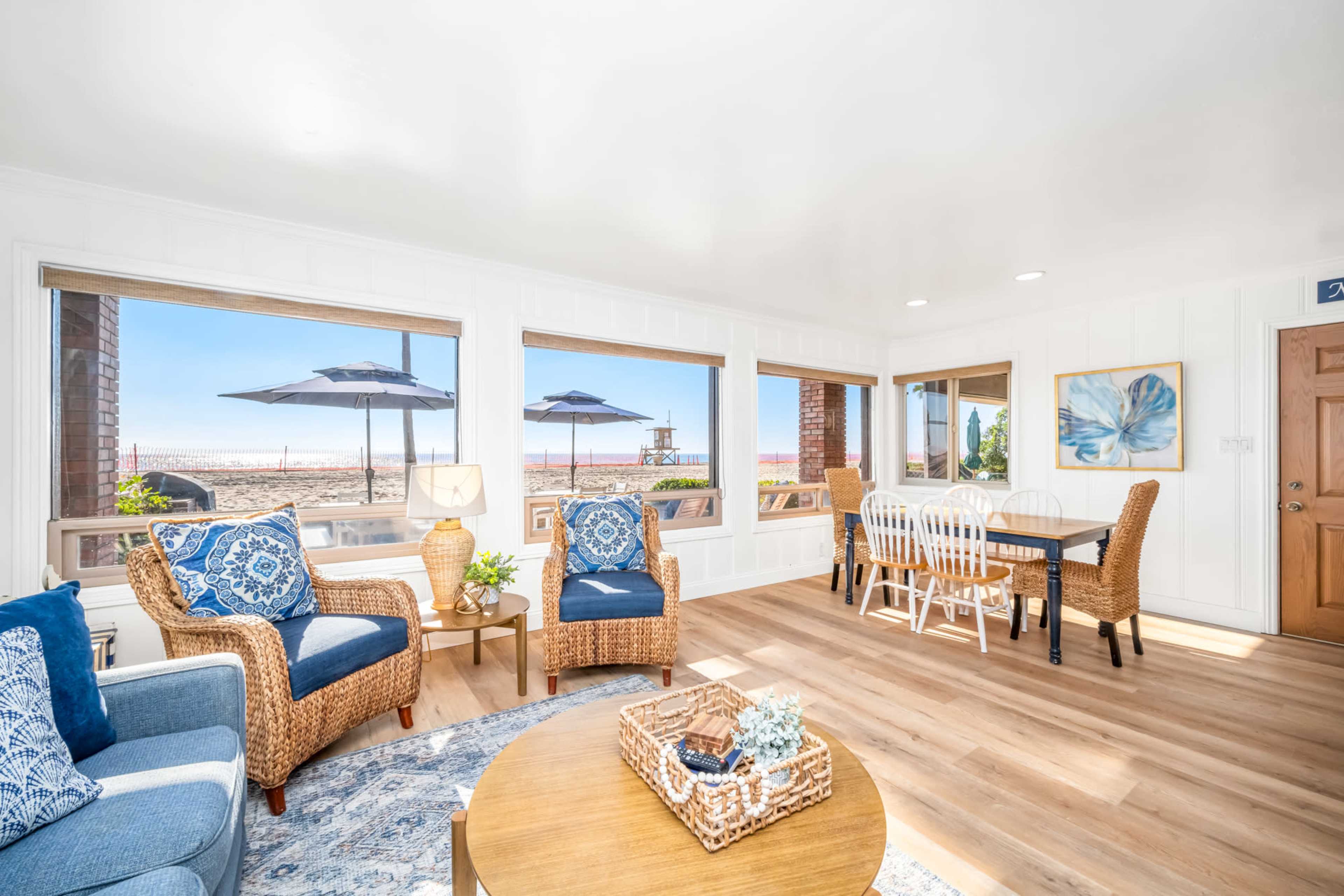 A bright living space features woven furniture, a round wooden table, and large windows showcasing a beach view with umbrellas outside.
