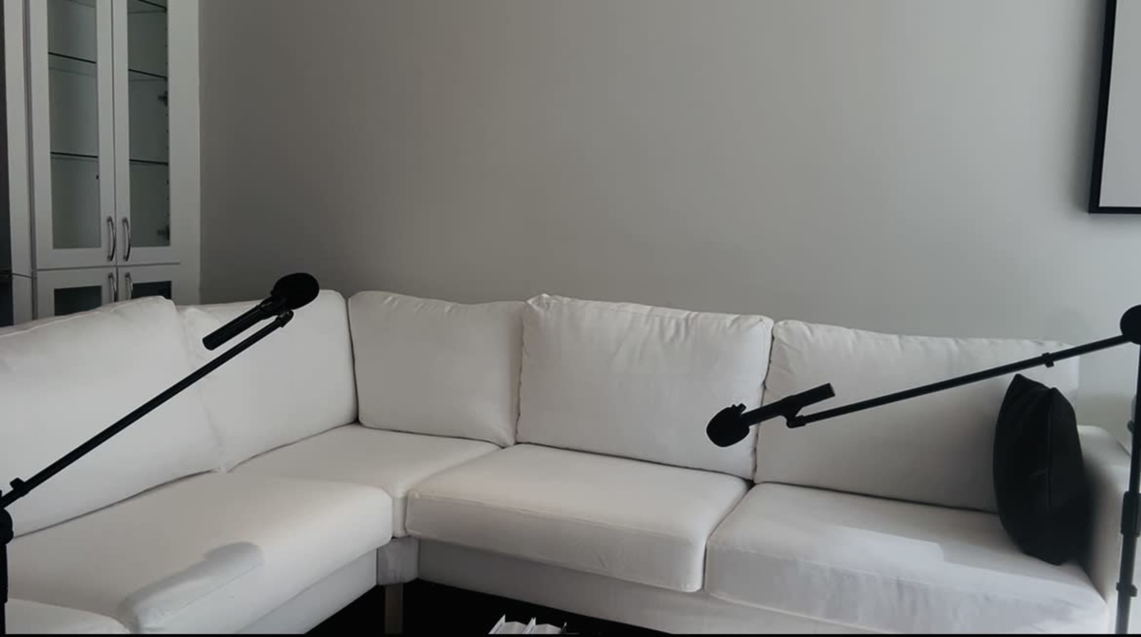 The image shows a white sectional couch in a minimalist room with two black microphones positioned towards it.