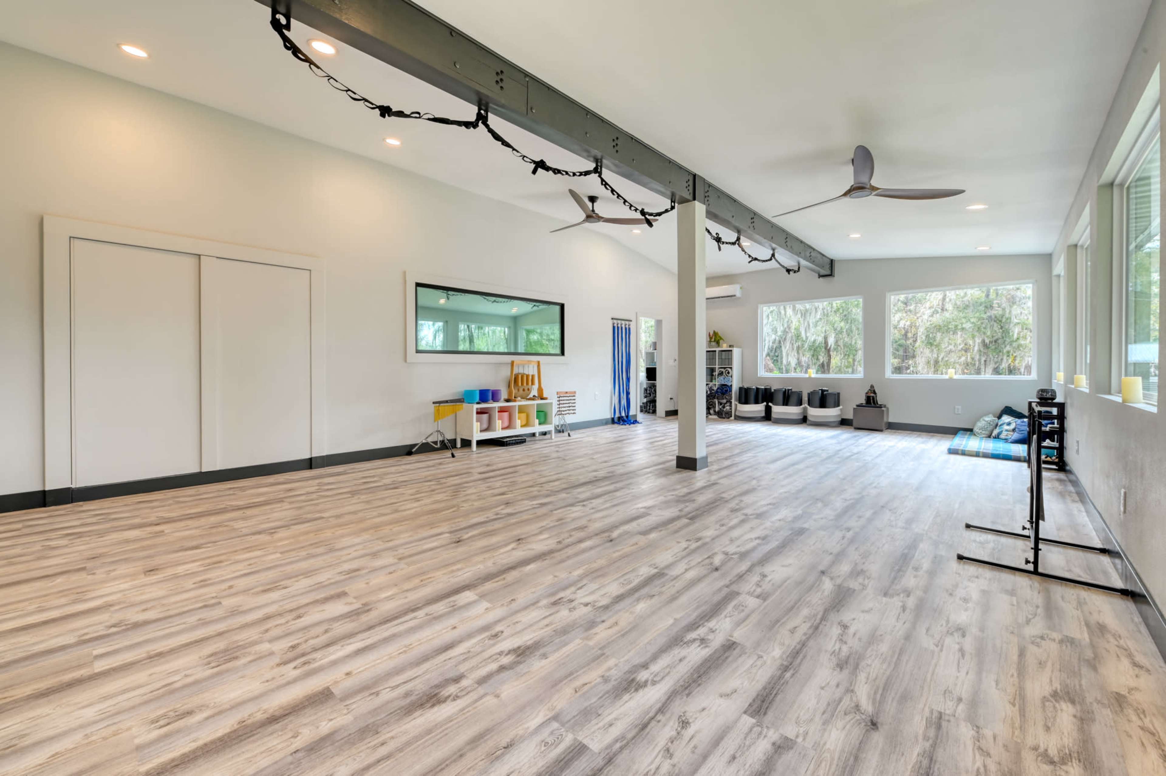 The image shows an empty room with light-colored flooring, large windows letting in natural light, and minimal furnishings, including a few exercise mats and storage bins.