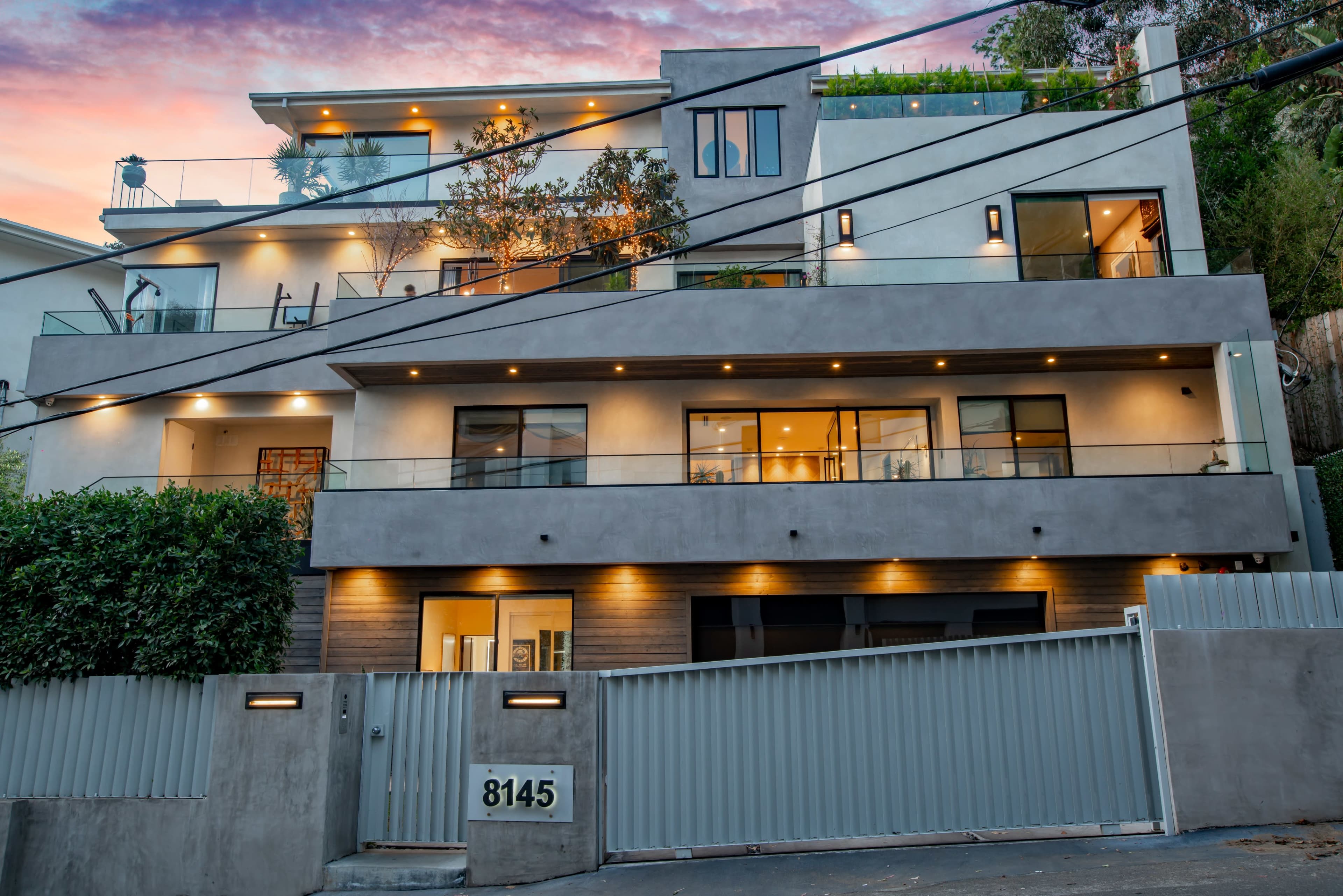 The image shows a modern multi-story house with large windows and a landscaped front yard, illuminated by outdoor lighting.