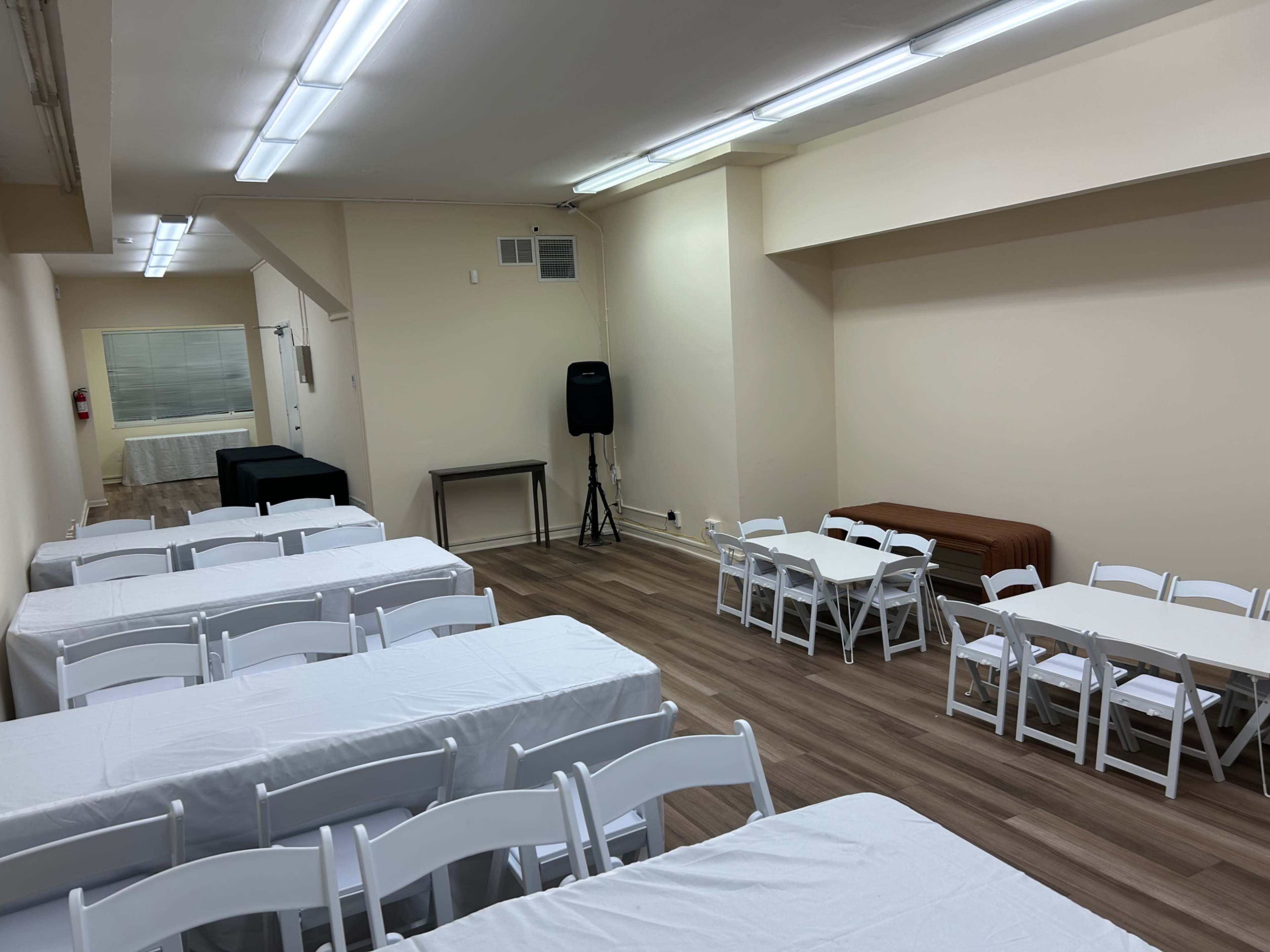 An empty room with white tables and chairs arranged in rows and a small stage area in the corner.