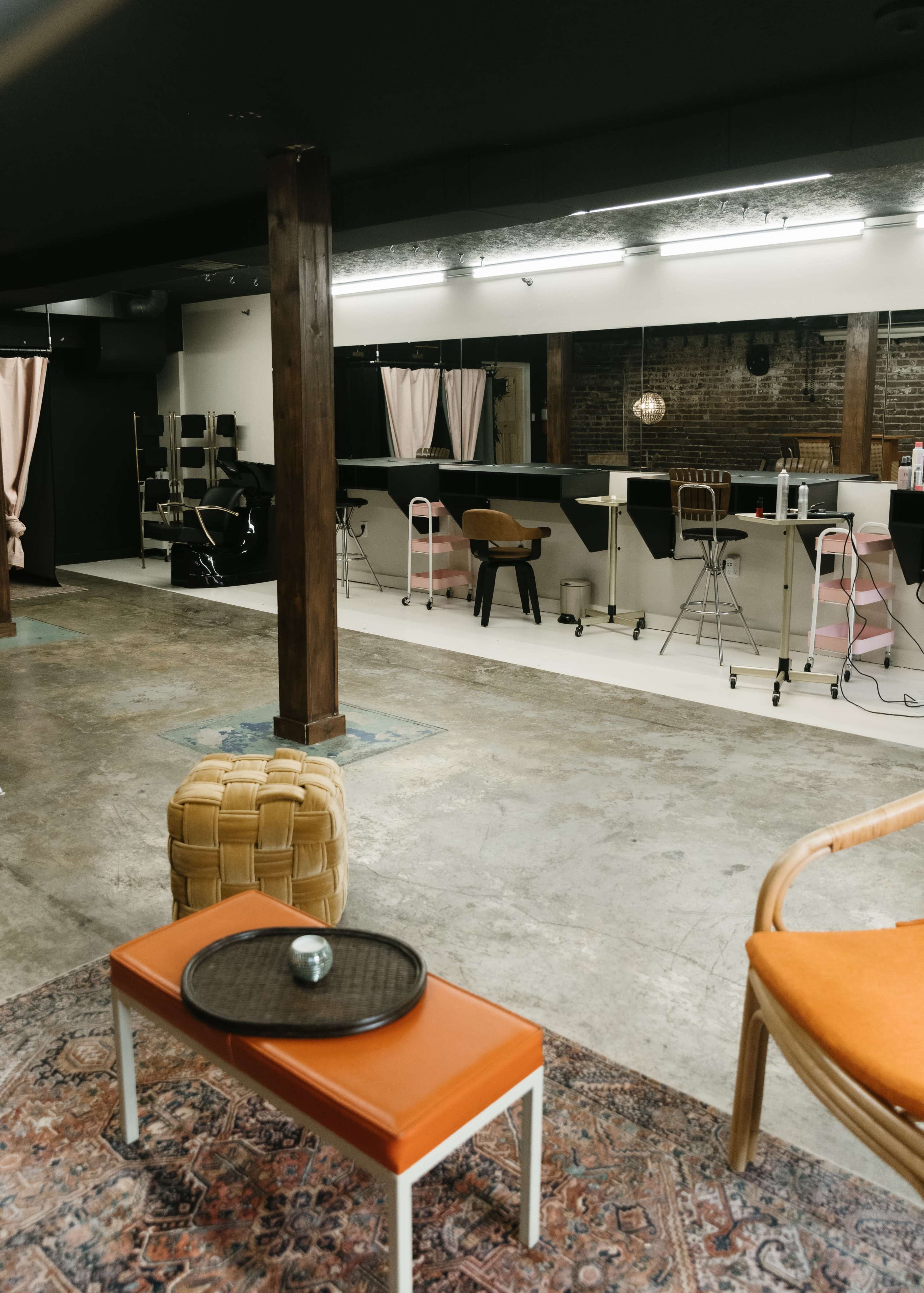 The image shows a hair salon interior with a concrete floor, a few styling stations, and seating areas featuring a wooden column and a patterned rug.