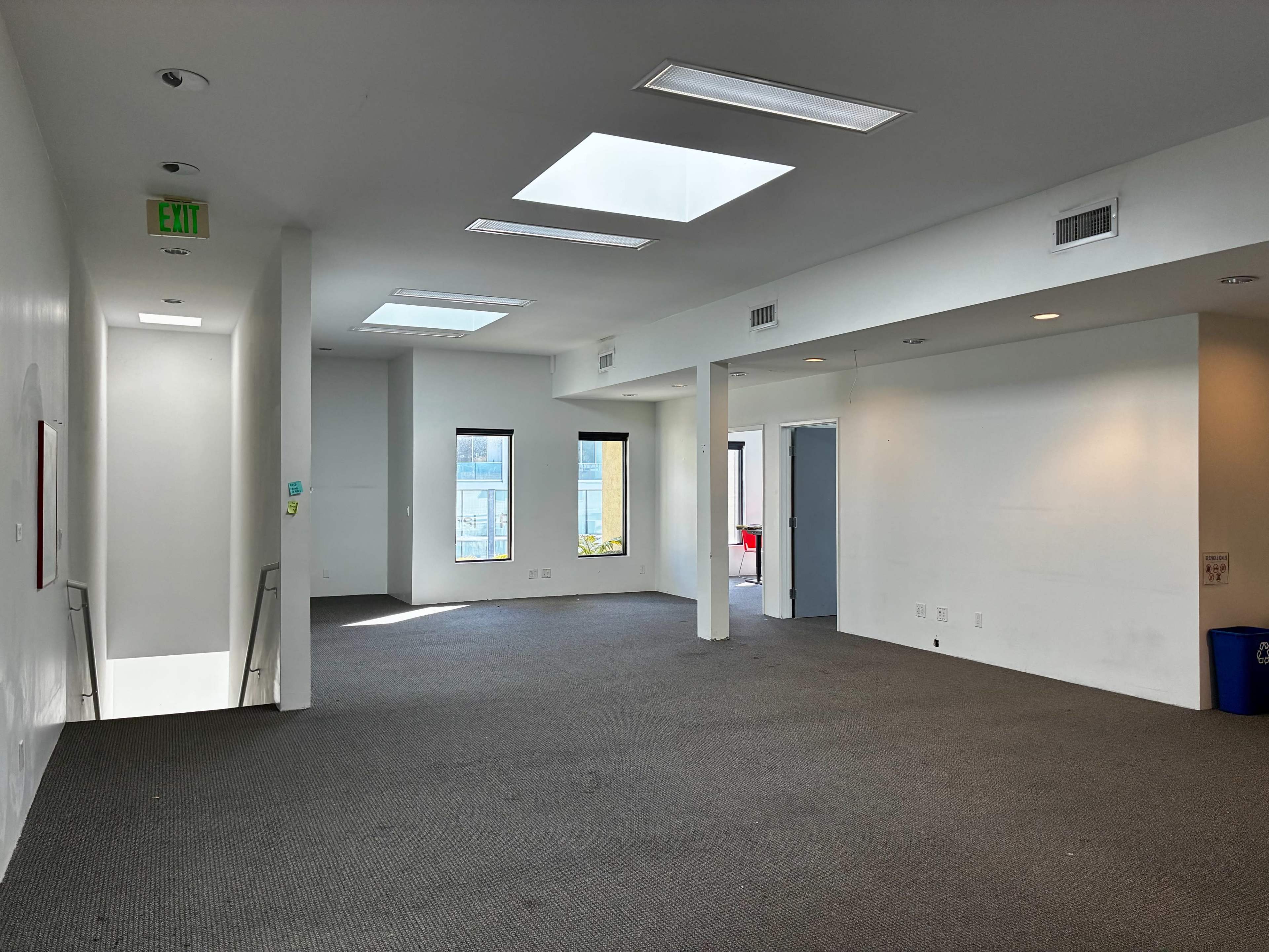 The image shows an empty room with gray carpet, large windows, and multiple skylights.