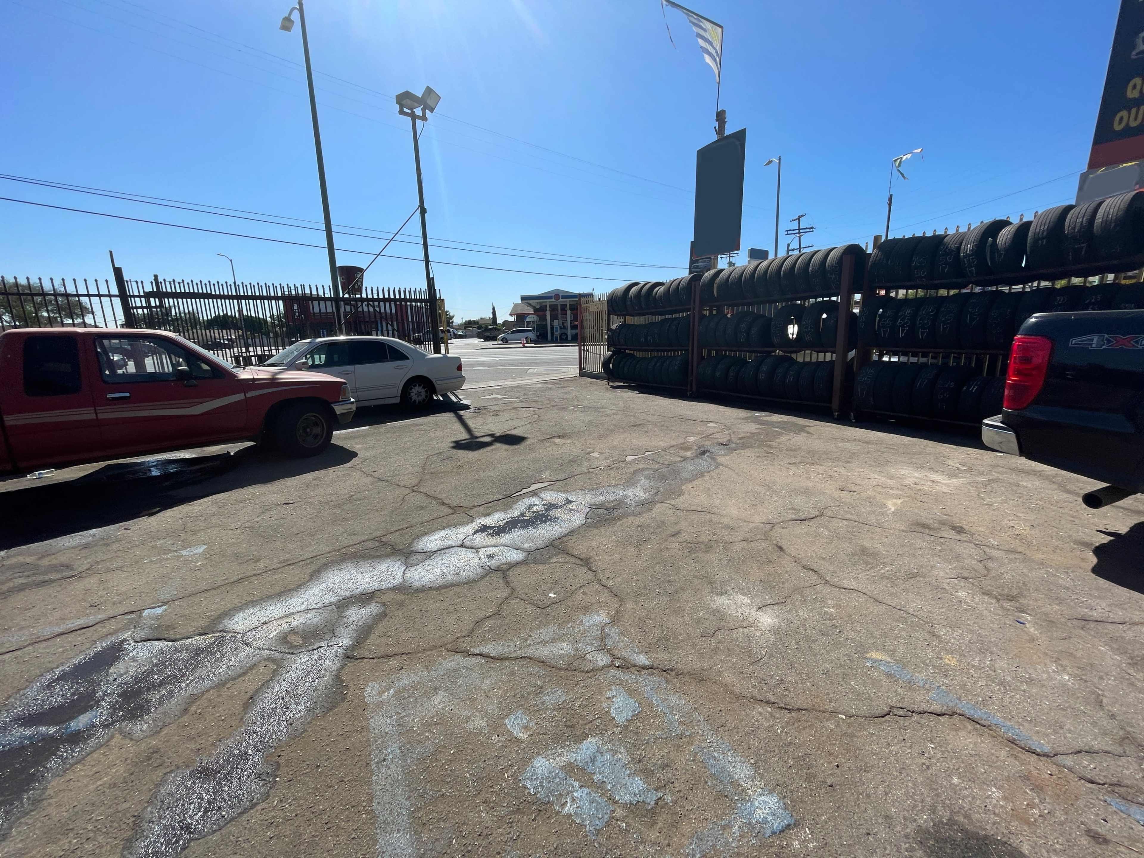 The image shows a parking lot with two vehicles, one red and one white, near a stack of tires along a fence under a clear sky.