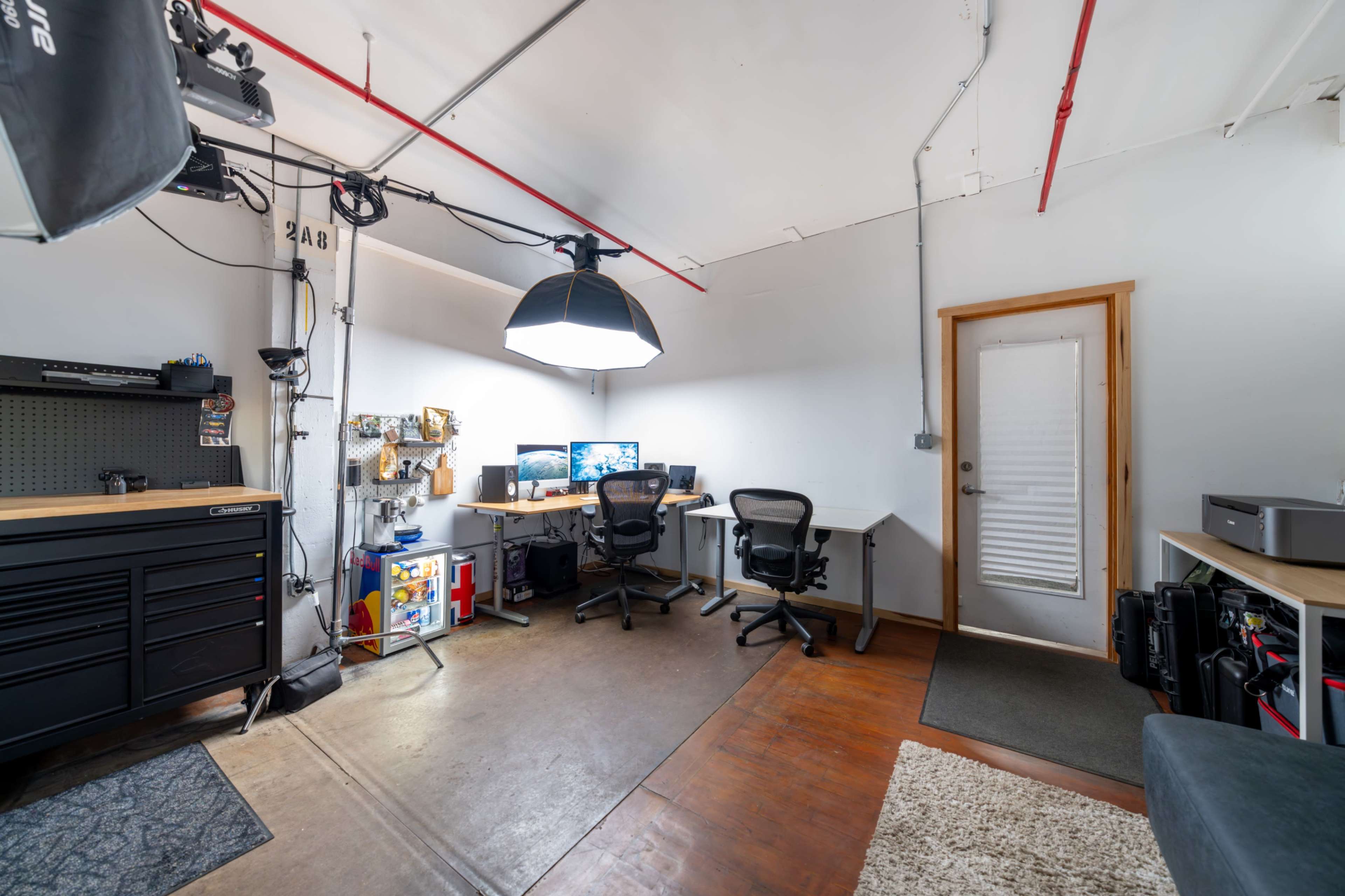The image shows a well-lit office space featuring two computer desks with chairs, a tool cabinet, and a door leading to an exterior area.