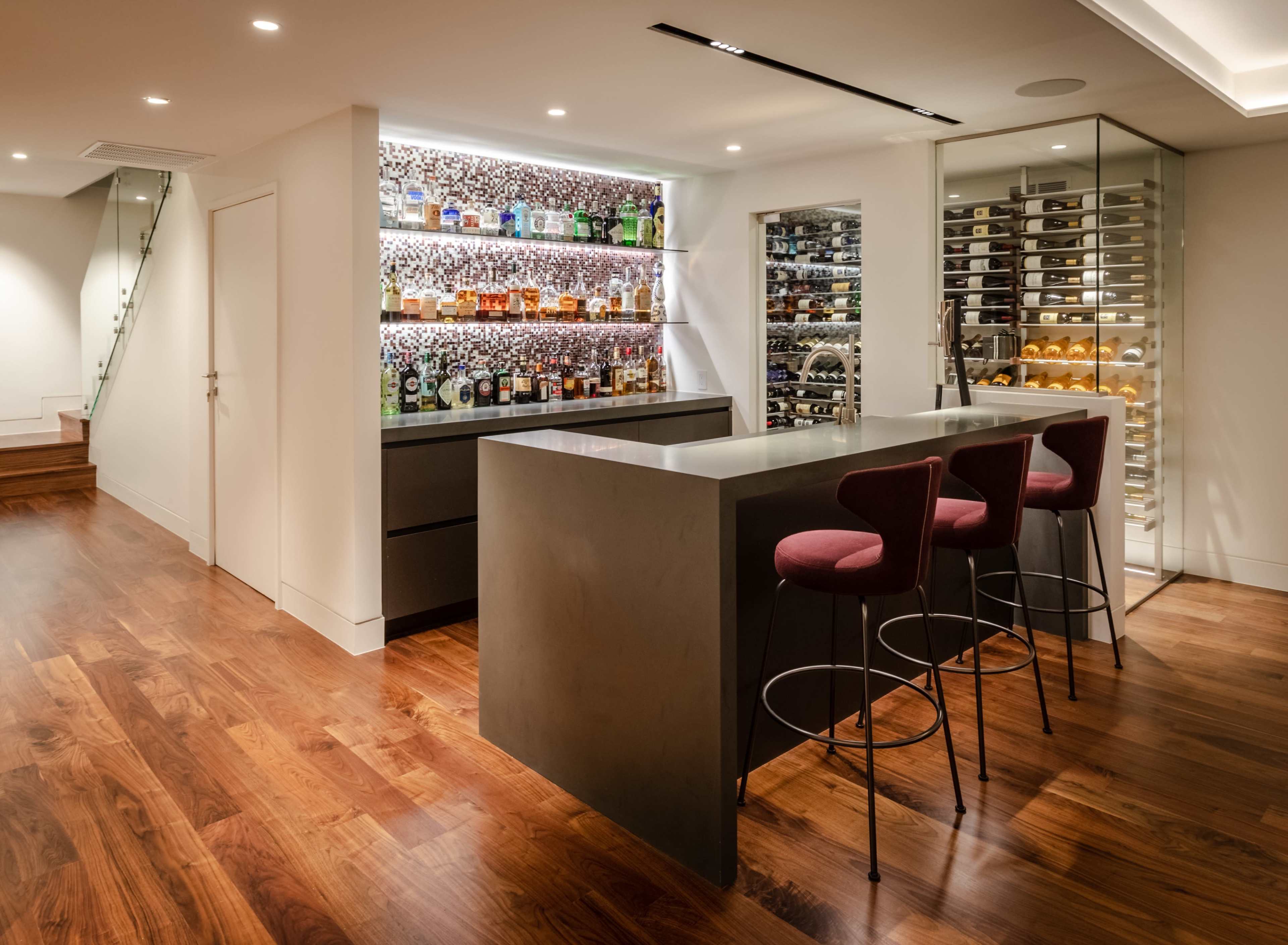 The image shows a modern home bar area with a sleek gray countertop, three red bar stools, and shelves displaying various bottles of liquor, alongside a wine fridge.