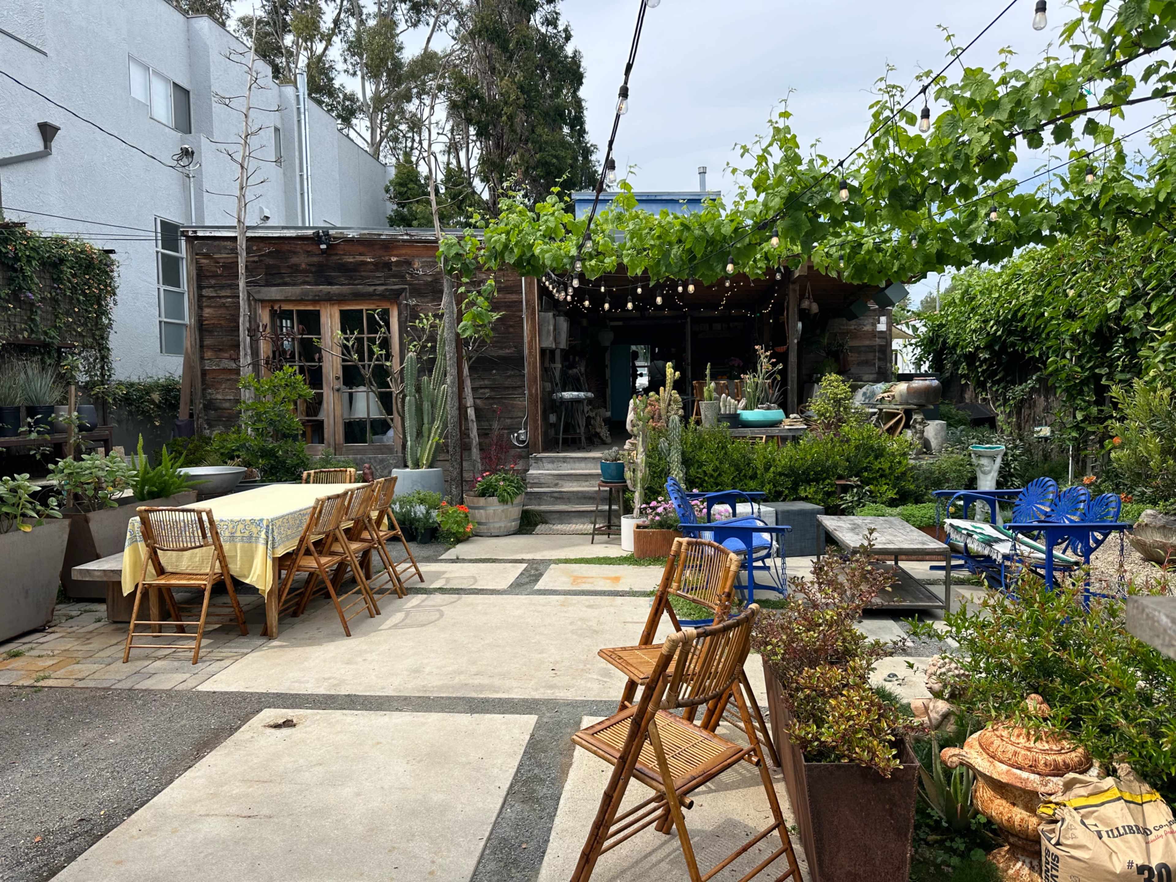 The image shows a landscaped outdoor garden area with wooden furniture, decorative plants, and a rustic building in the background.