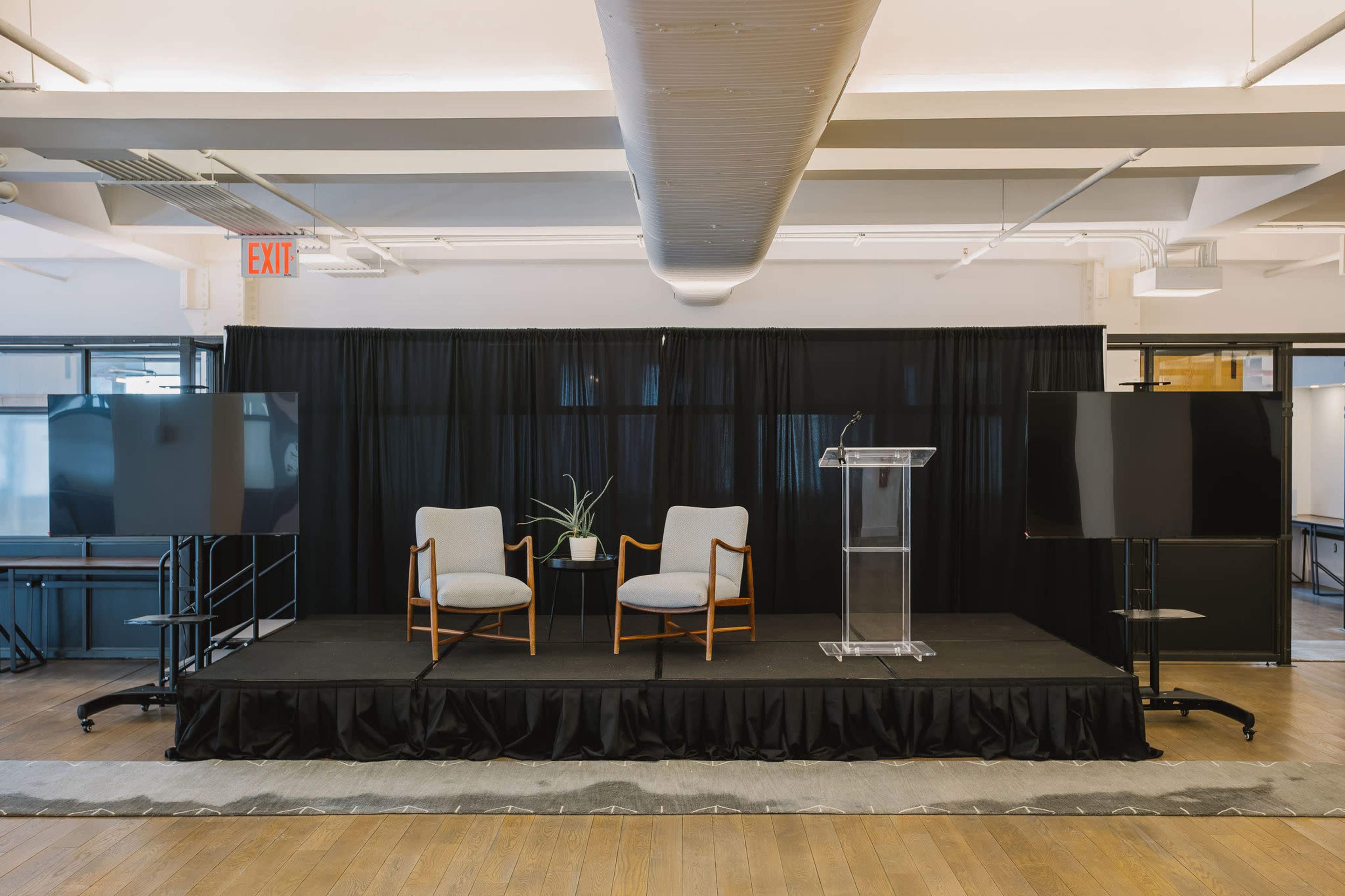 The image shows a stage setup with two chairs and a small table accompanied by a podium, all in front of a black curtain, with two large screens on either side.