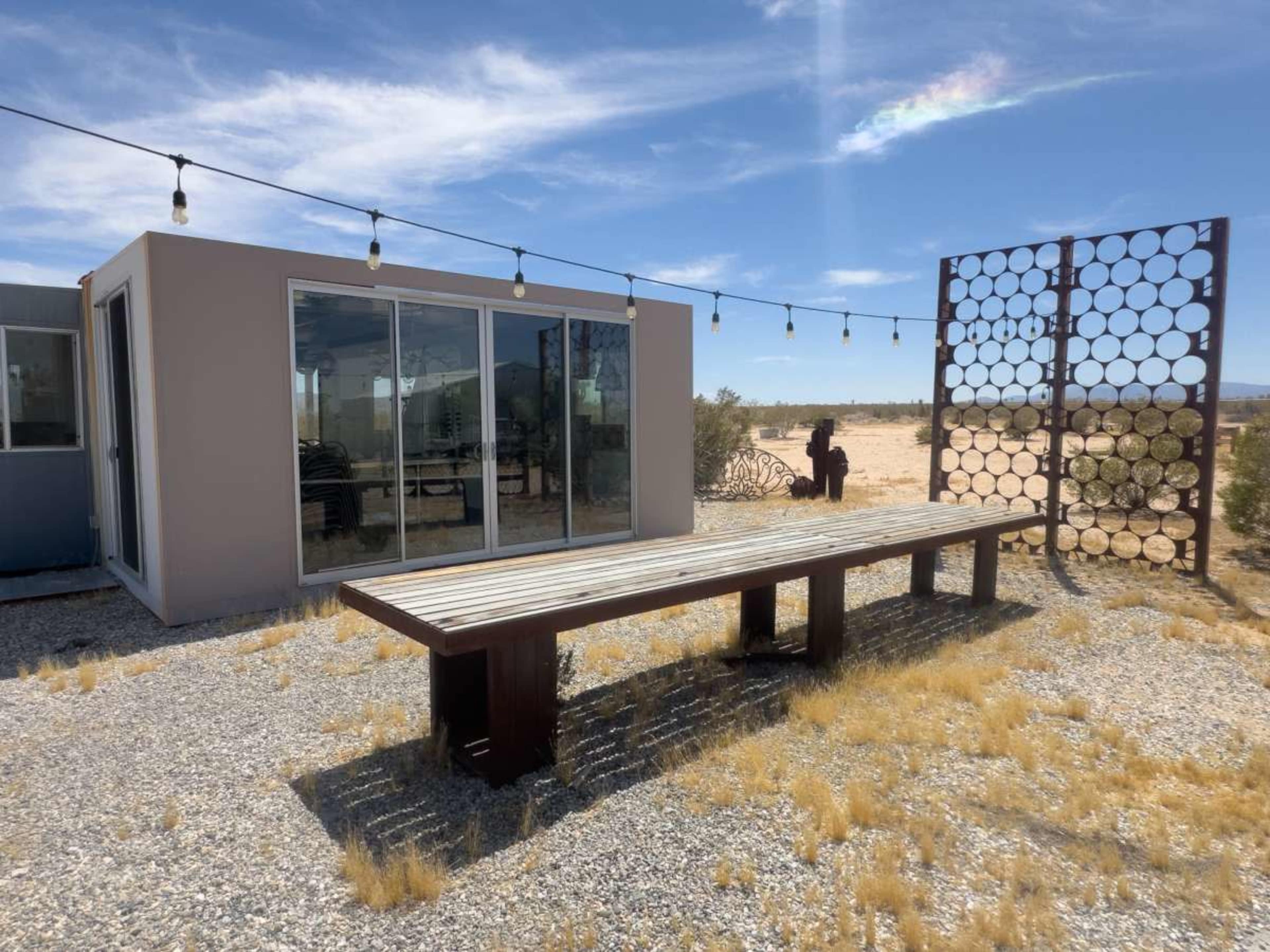 A modern building with large glass windows is situated beside a long wooden table in a desert landscape, with a decorative screen and string lights overhead.