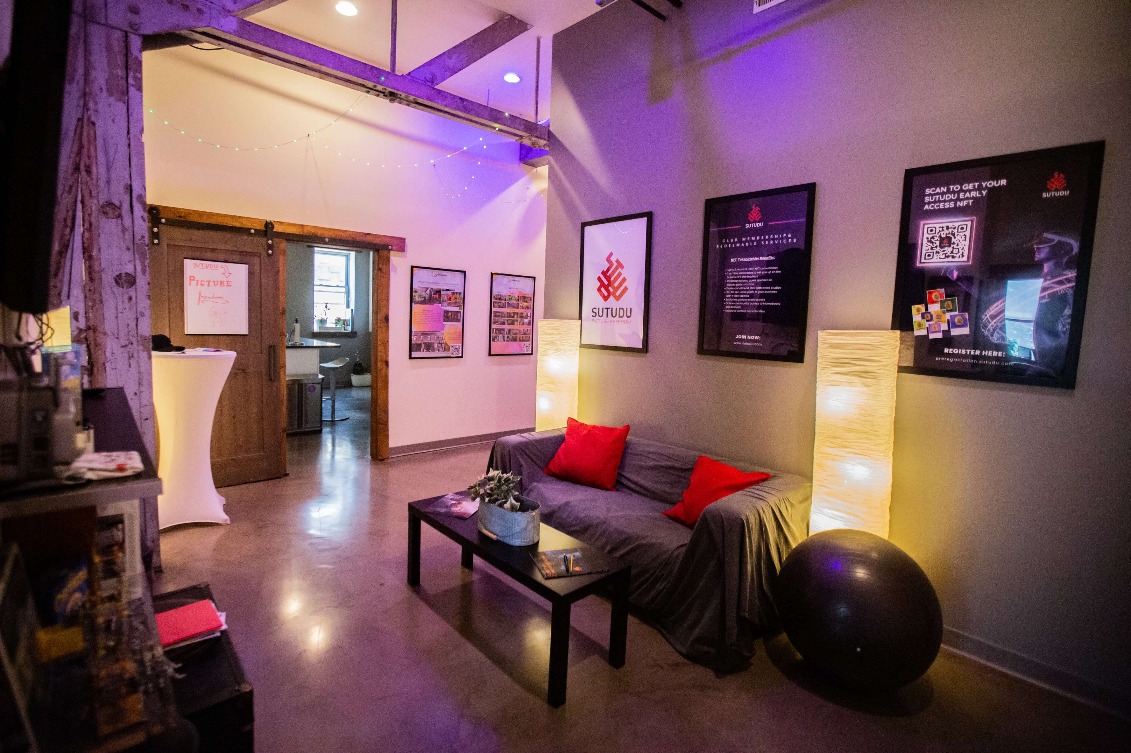 A cozy lounge area with a gray sofa, red accent pillows, decorative floor lamps, and framed posters on the walls.