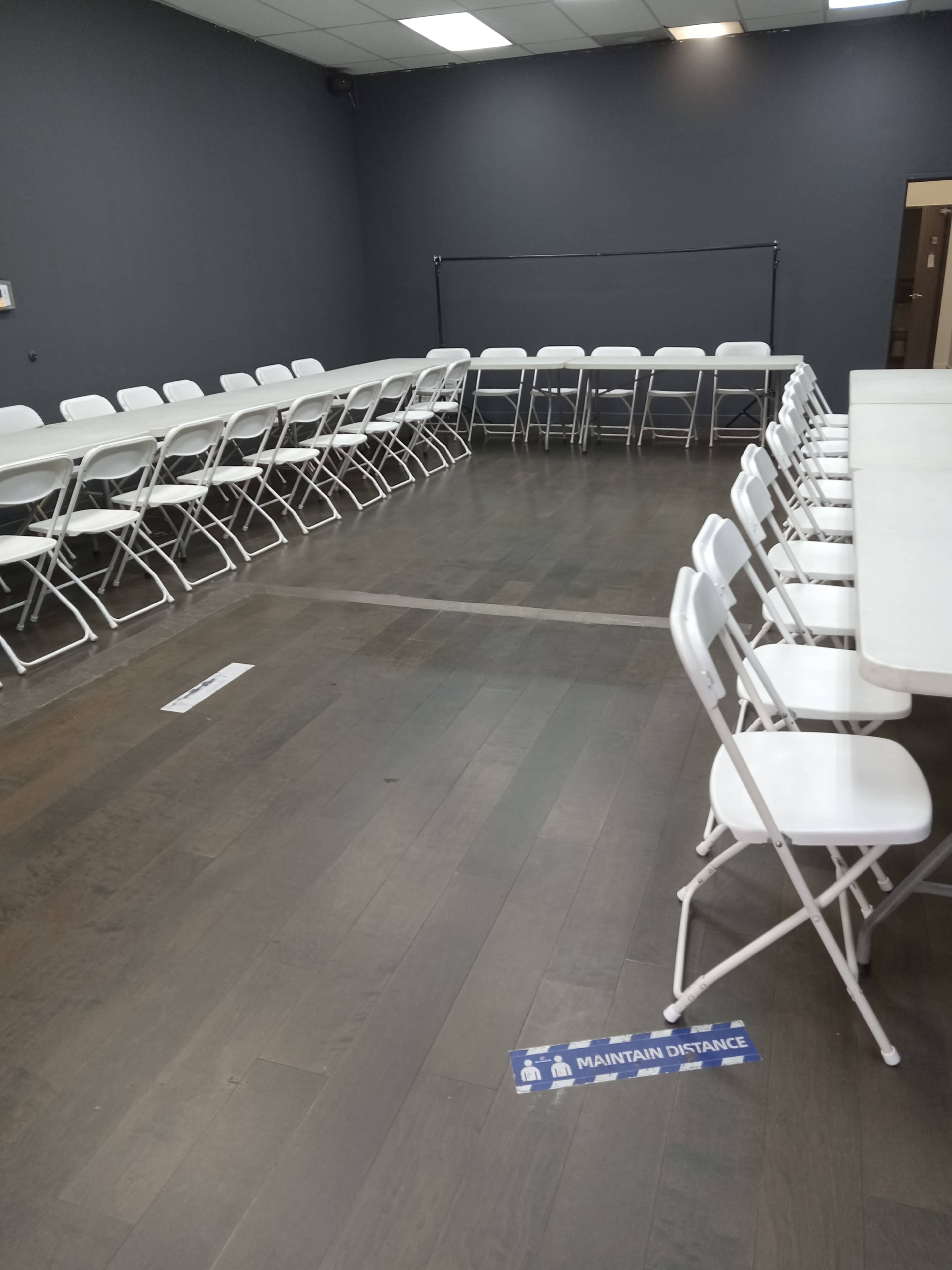 The image shows a sparsely arranged meeting room with white folding tables and chairs set up in two rows along the walls, as well as floor markings indicating to maintain distance.