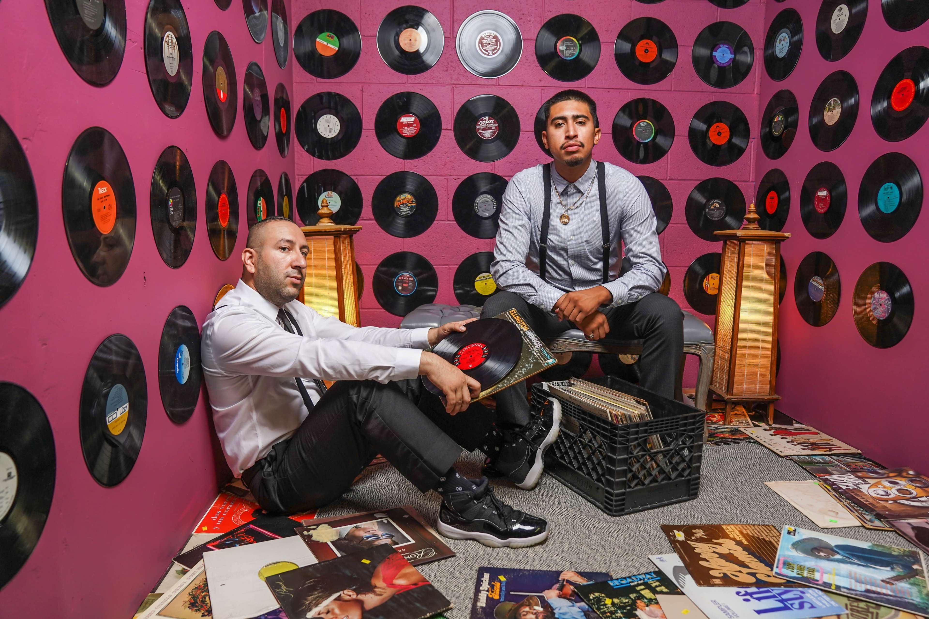 Two men sit among scattered vinyl records on the floor, surrounded by a wall adorned with vintage records, while one man examines an album and the other poses thoughtfully.