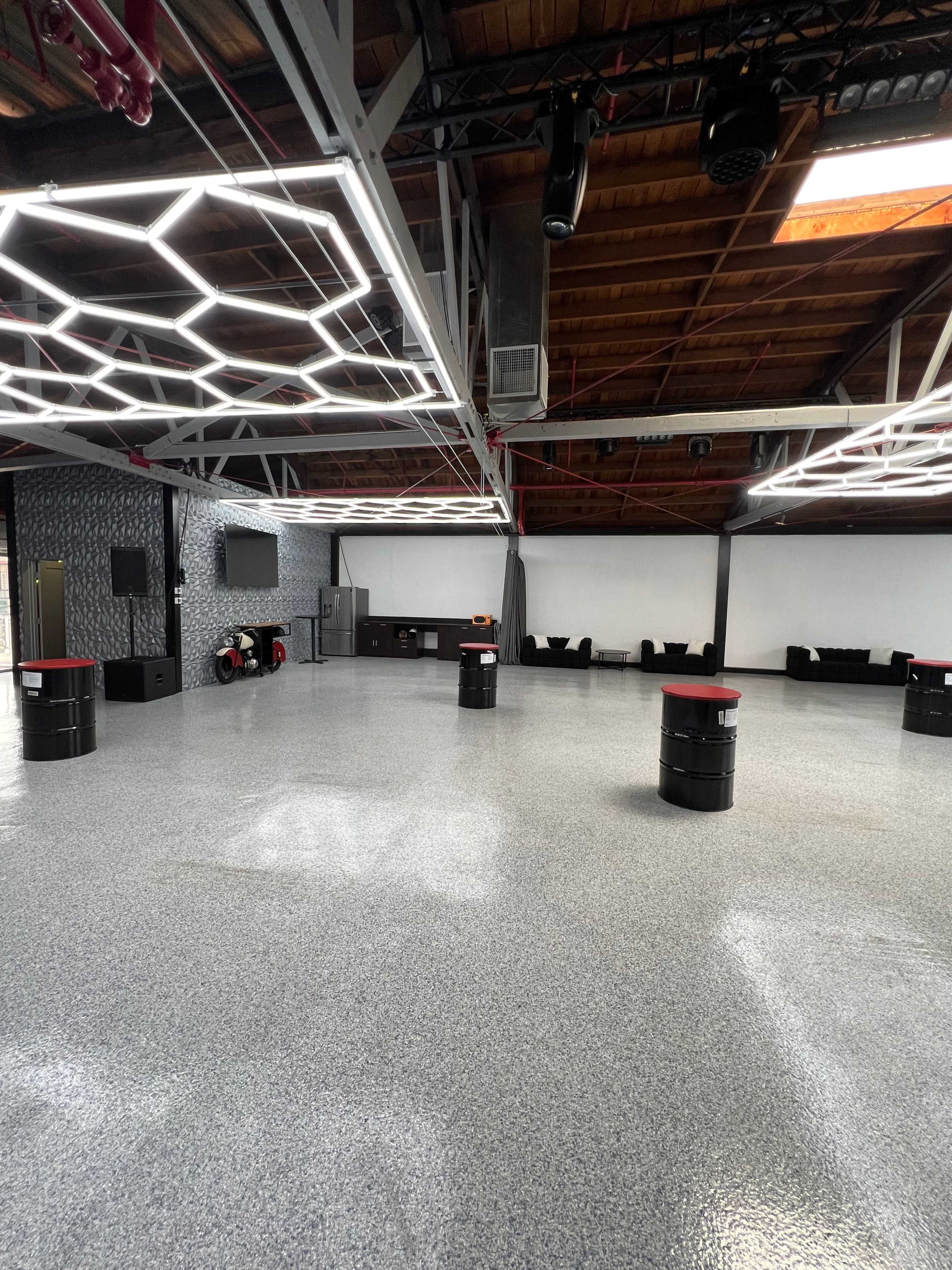 A spacious interior room with a polished floor, modern lighting fixtures, and a mix of seating areas and tables made from black barrels.