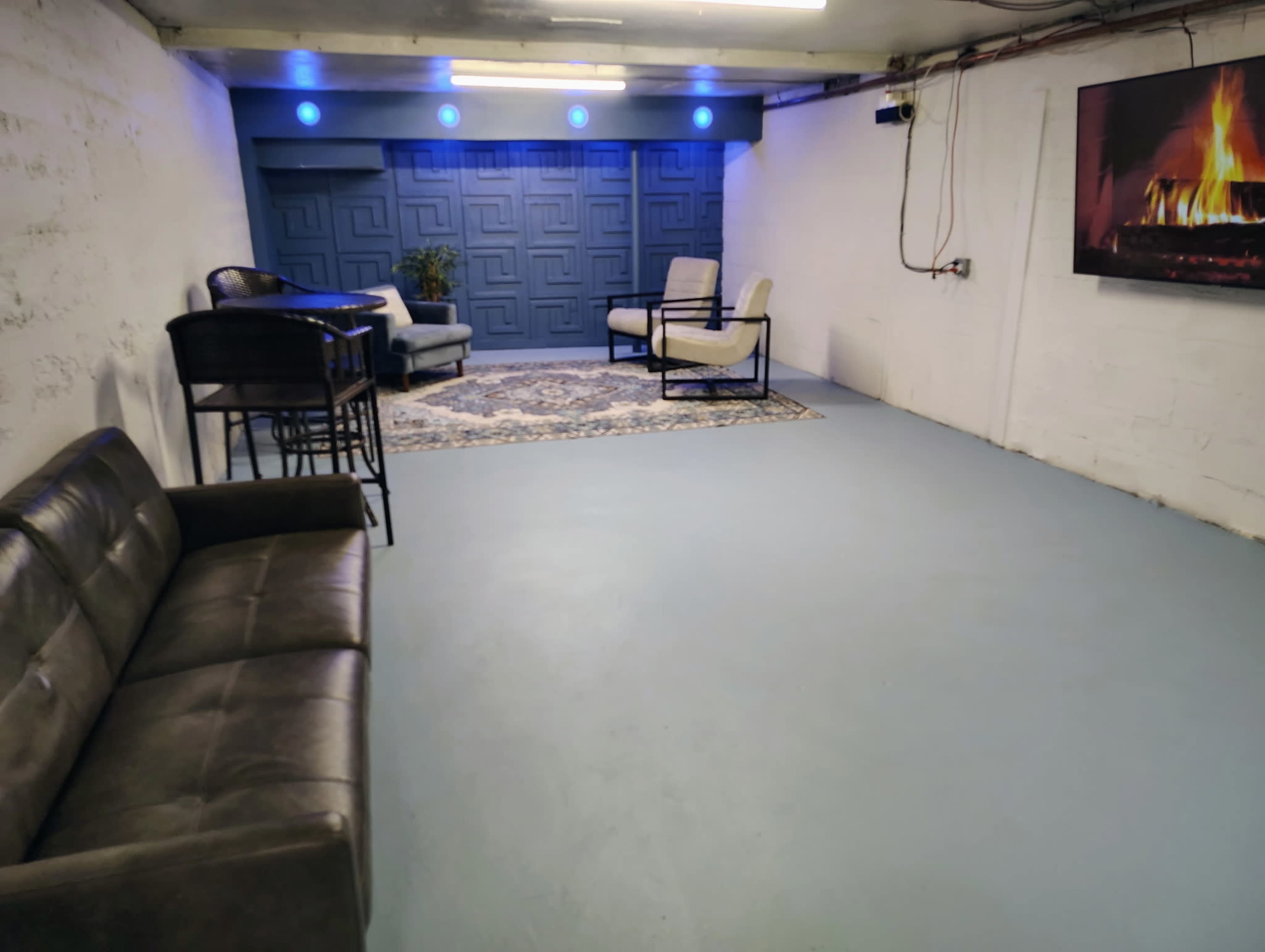The image shows a spacious basement area with a blue floor, a couch, two chairs, a small table, and a wall-mounted television displaying a fireplace.