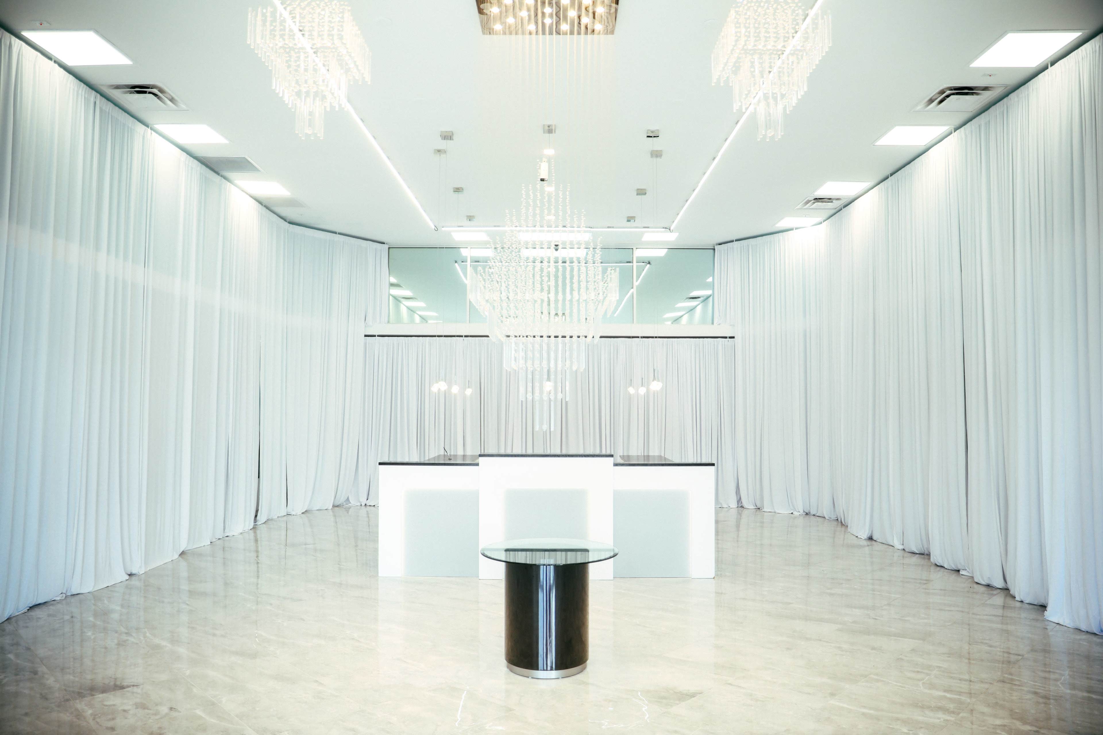 The image shows an empty event space featuring white curtain drapery, two large chandeliers, and a central round table.