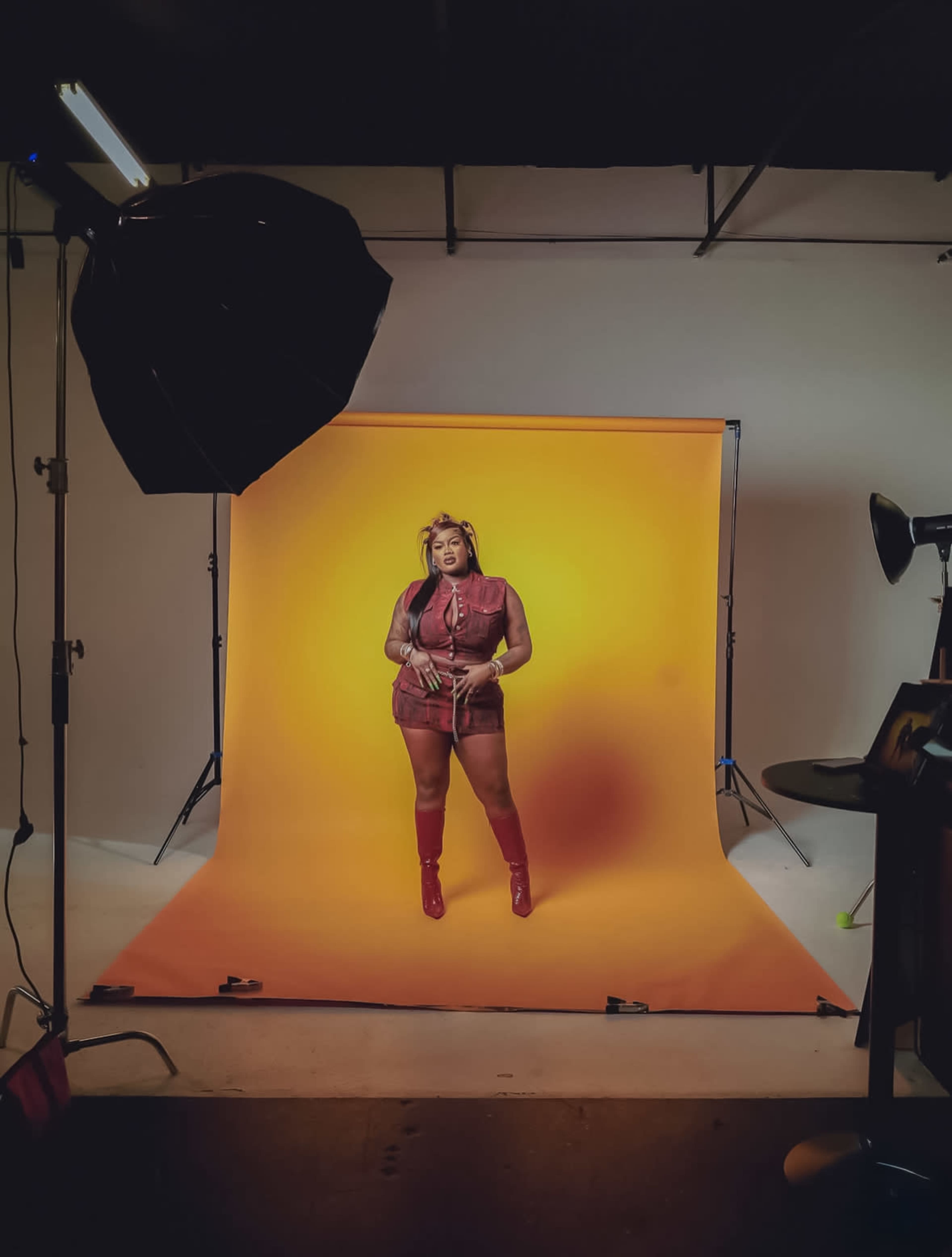 A model poses against a vibrant orange backdrop in a studio setting, illuminated by professional lighting equipment.