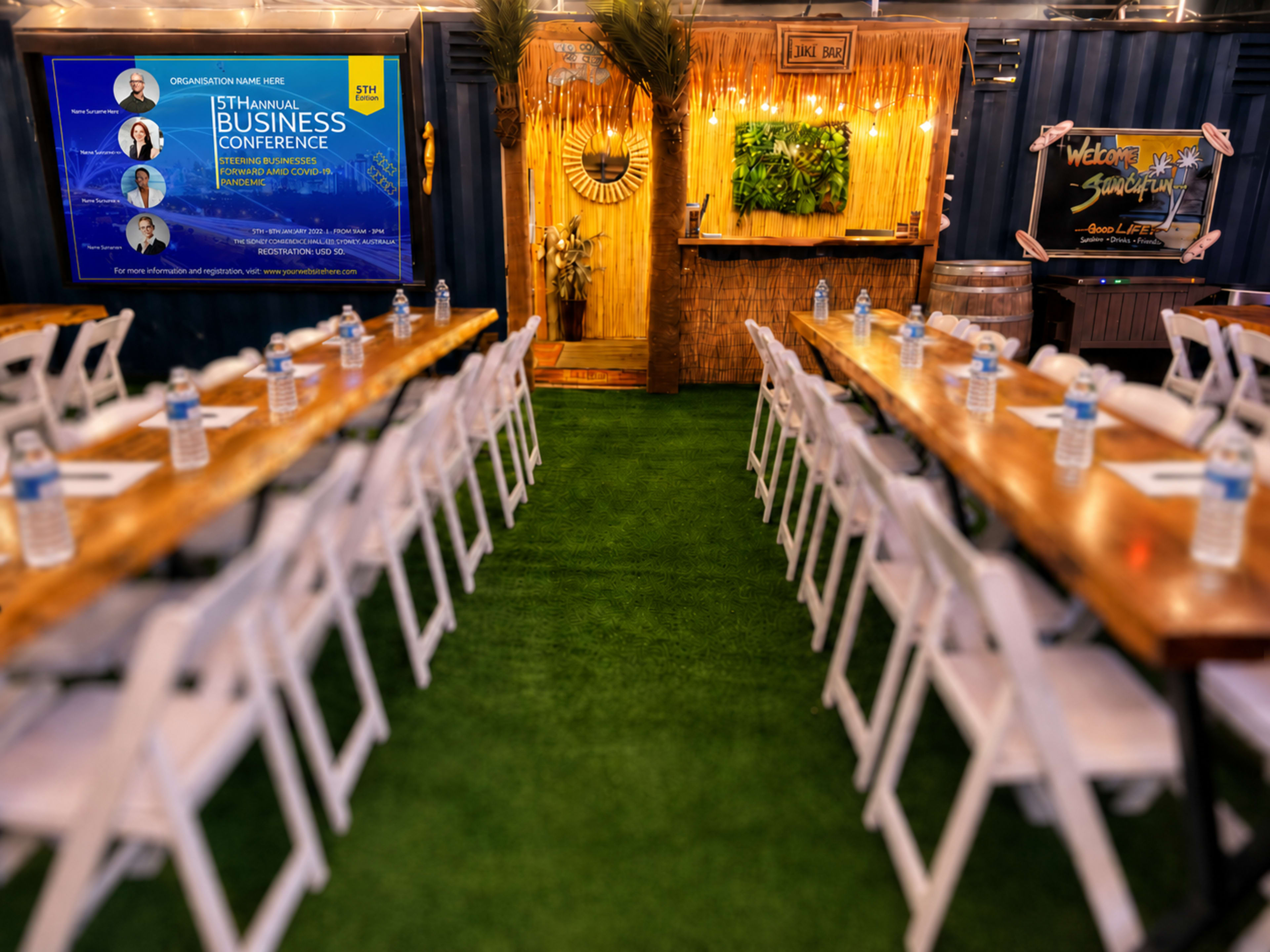 The image shows a conference setup with rows of wooden tables and chairs arranged on a green carpet, flanked by a blue presentation screen and tropical-themed decor.