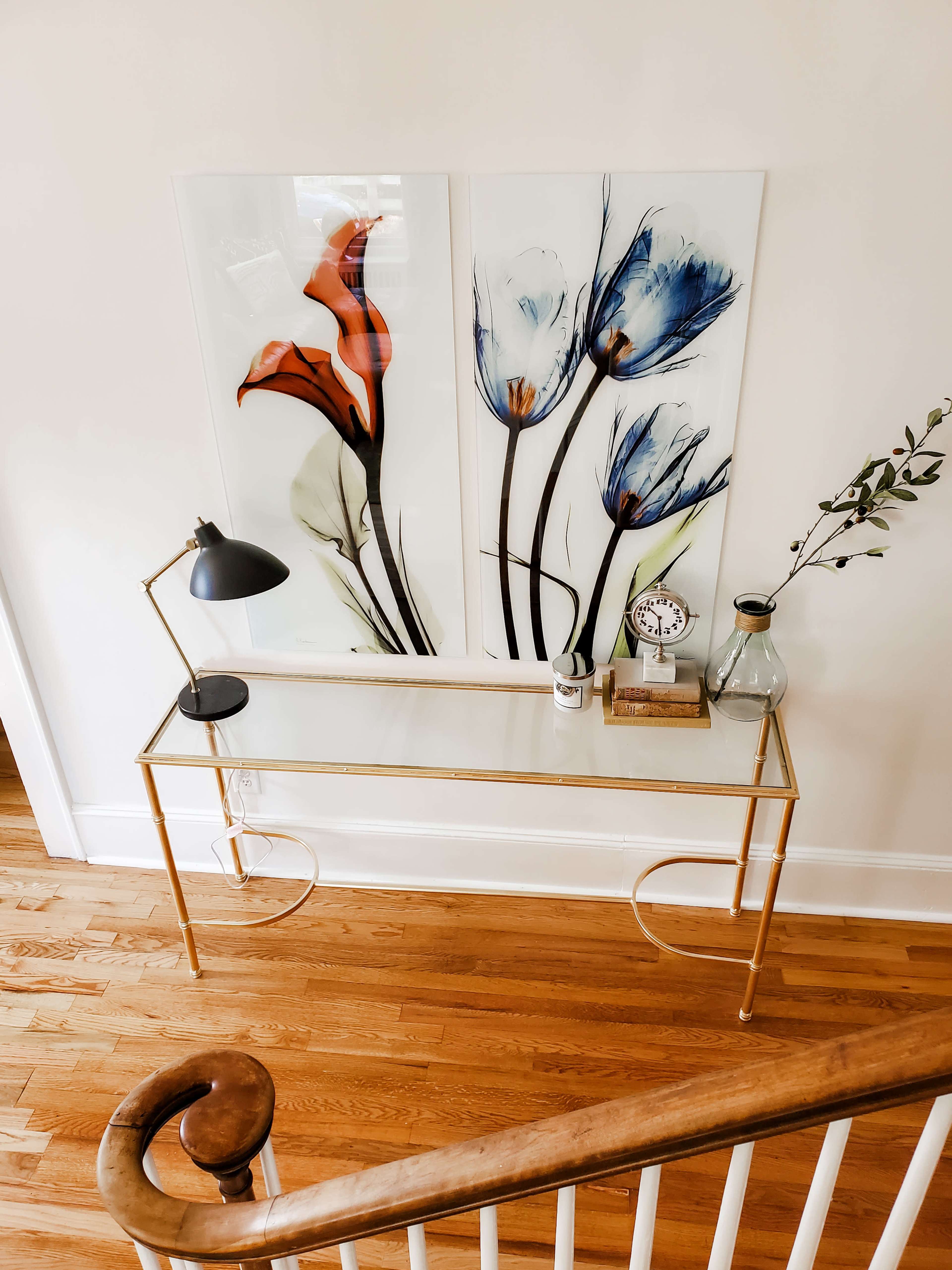 A gold metal console table with a glass top is displayed against a wall featuring two floral artworks, accompanied by a lamp, a clock, and a decorative vase.