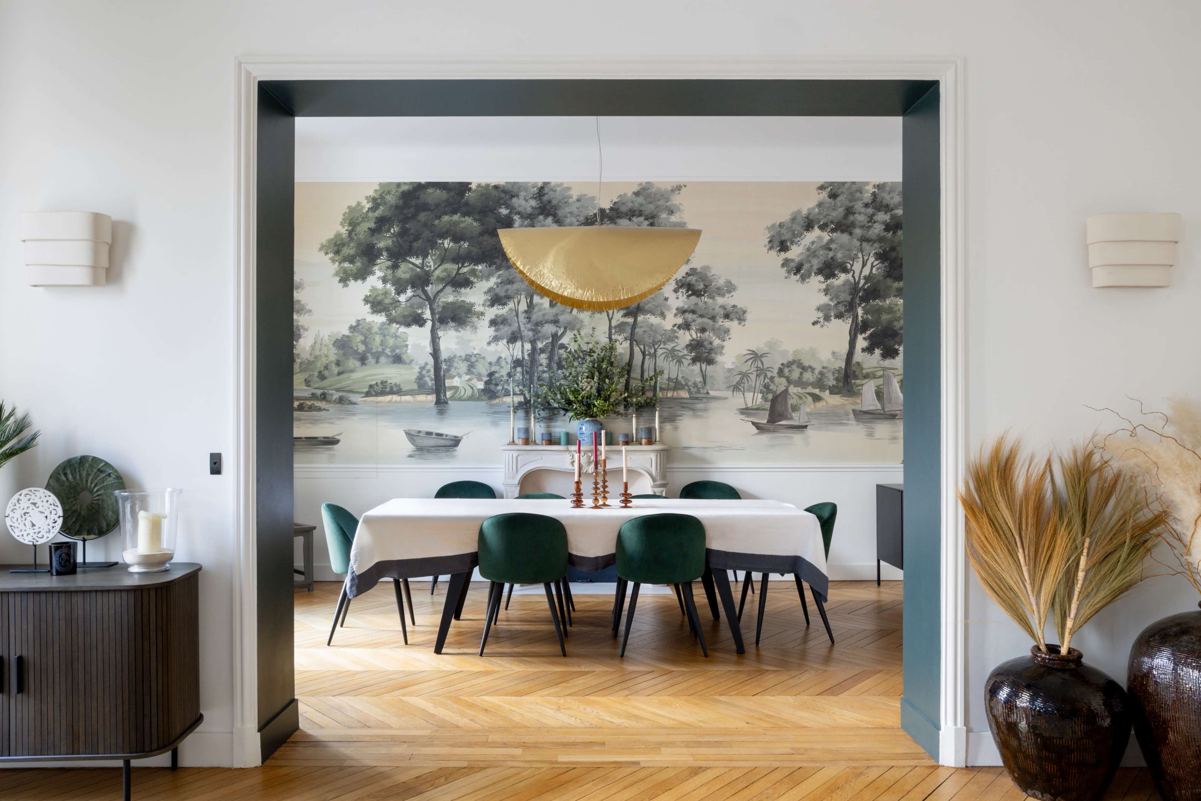 The image shows a dining room framed by an archway, featuring a large table covered with a white tablecloth, surrounded by green chairs, and a mural of boats and trees in the background.