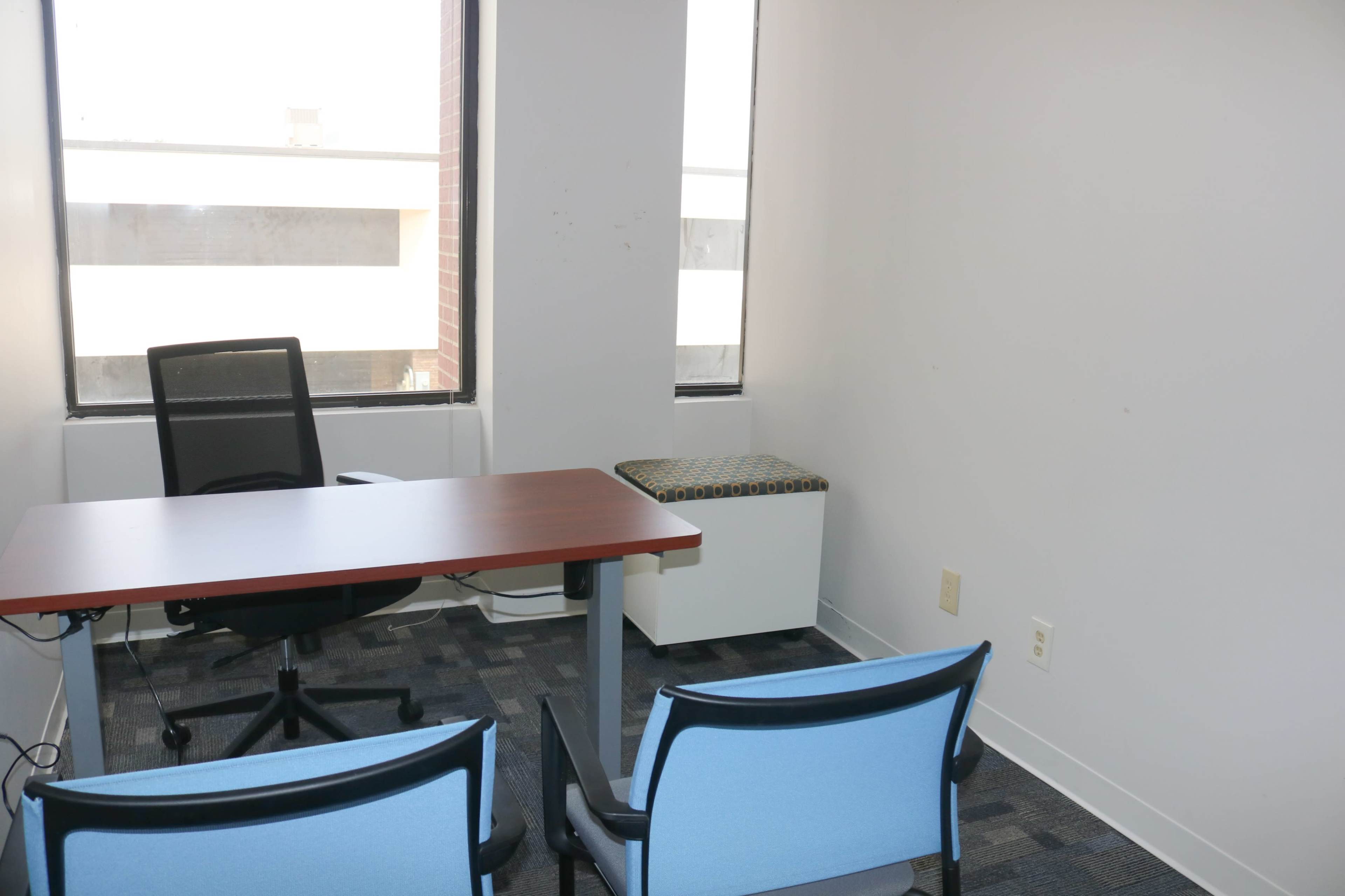 The image shows a small office space with a wooden desk, a black office chair, and two blue chairs facing the desk, along with a cushioned window seat.