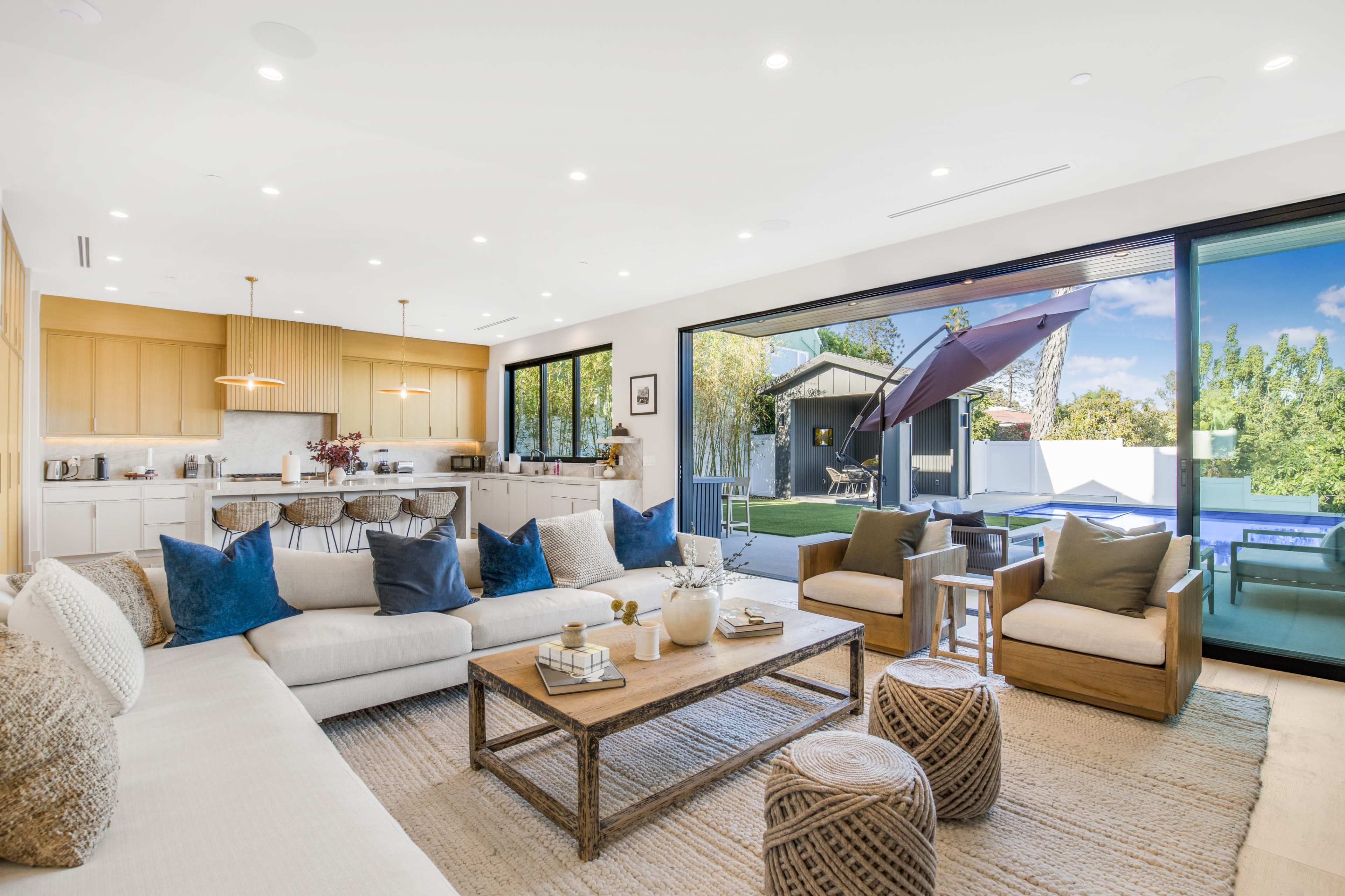 The image shows a modern living room with a spacious layout, featuring a light-colored sofa, wooden chairs, a coffee table, and large glass doors opening to a patio.
