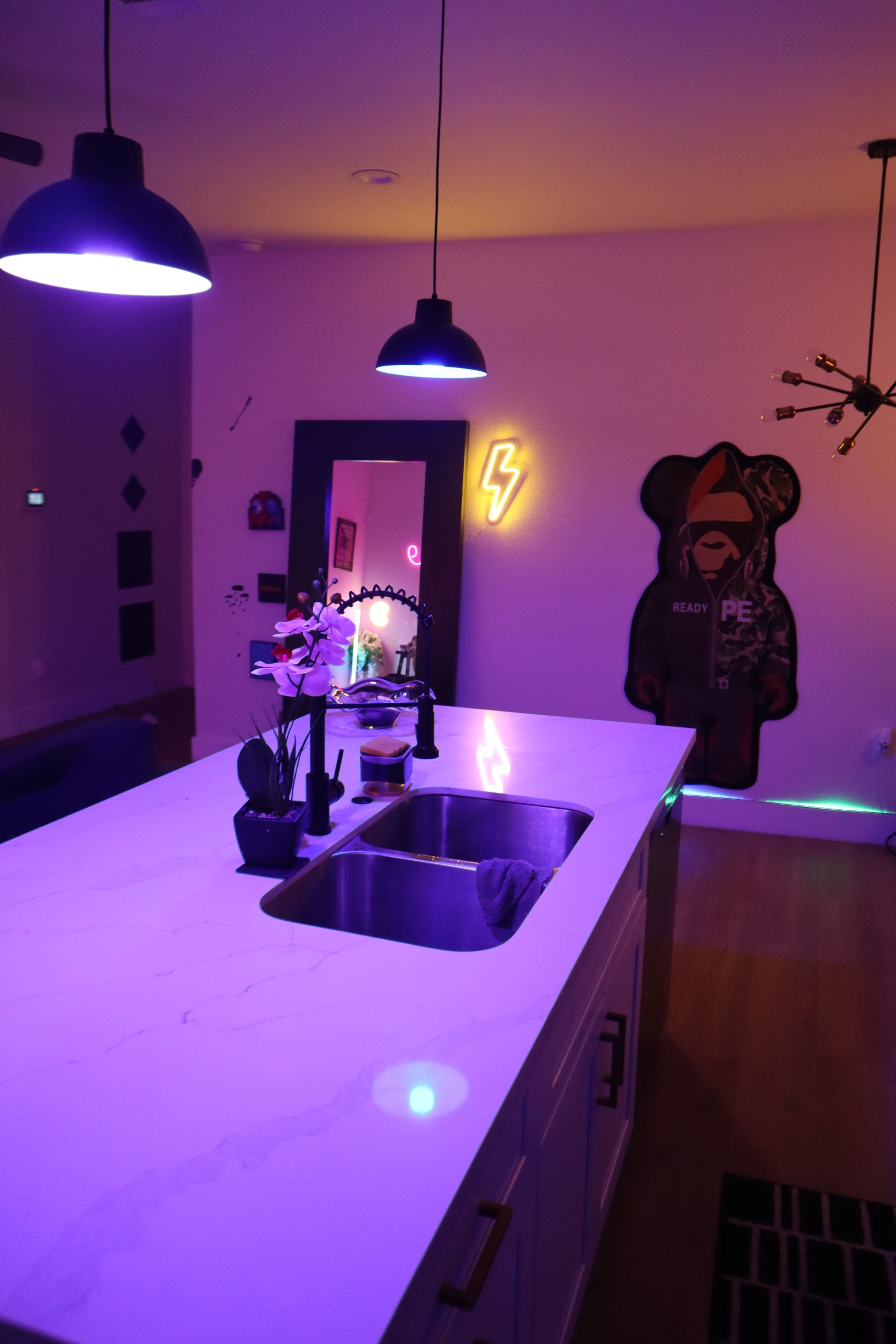 The image shows a modern kitchen with a white marble countertop, dark pendant lights, and neon decor on the walls, including a bear-shaped figure and a lightning symbol.