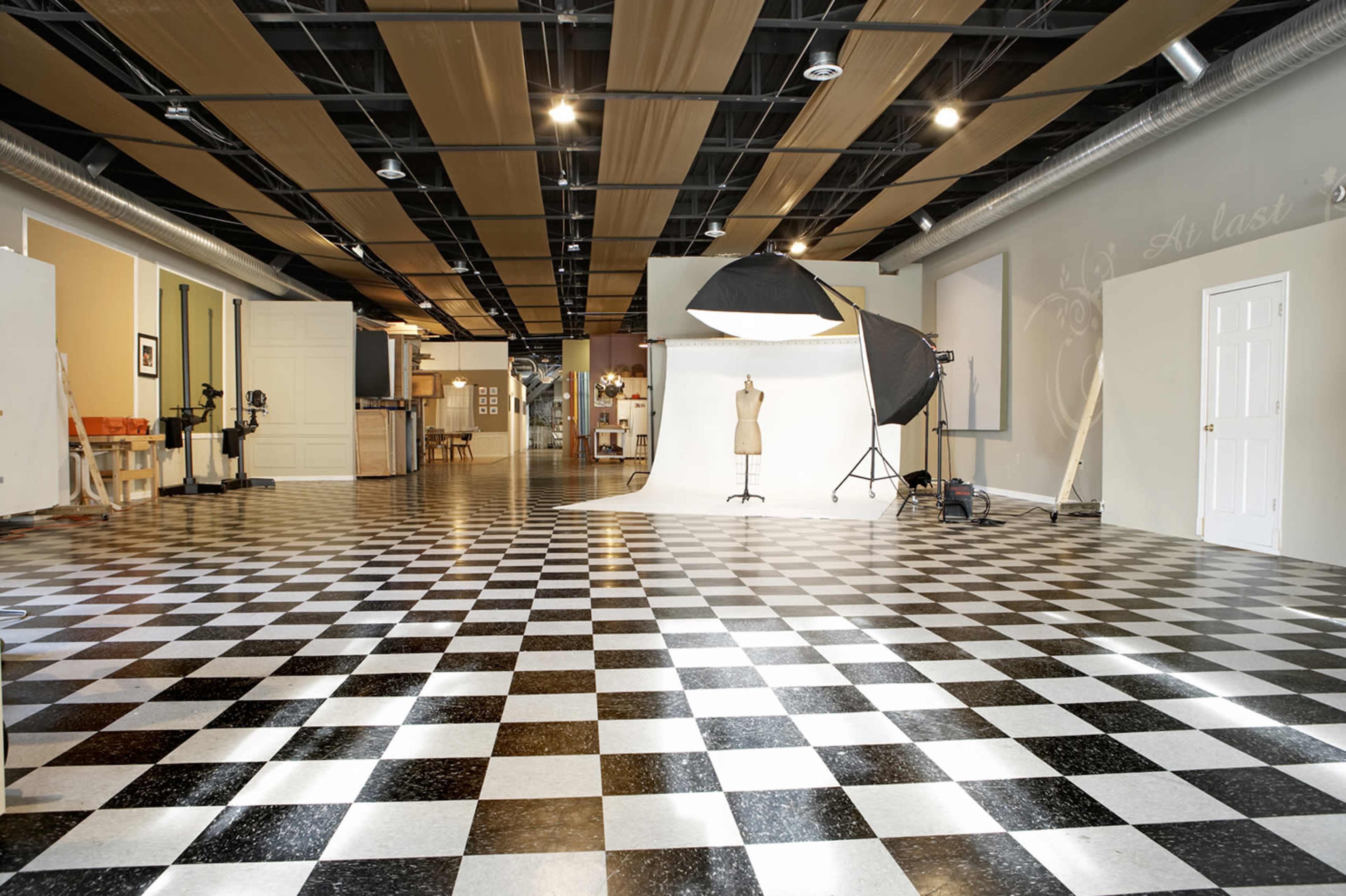 The image shows a spacious photography studio with a checkered floor, two white backdrop stands, a mannequin, and various equipment set up.