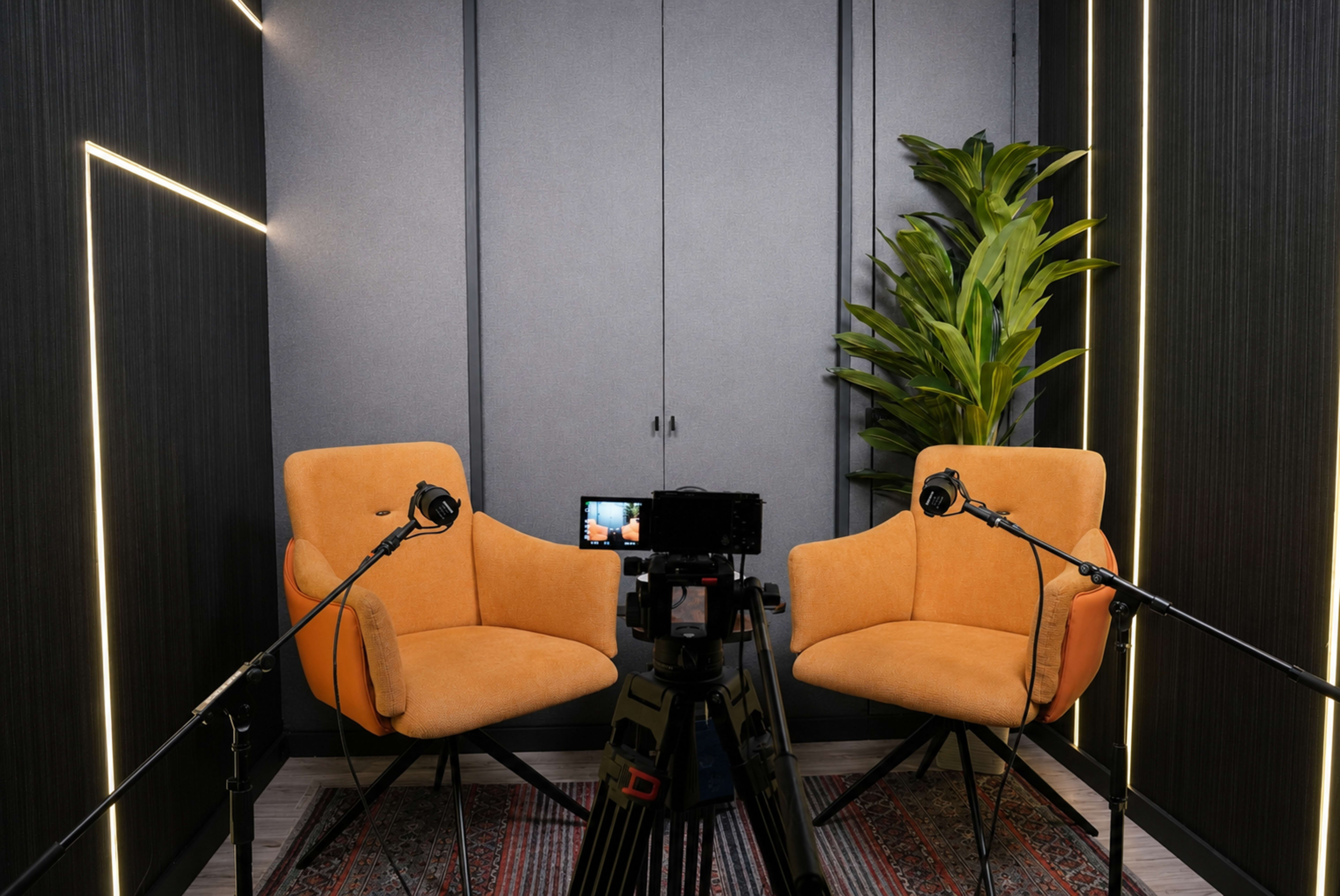A video recording setup features two orange chairs facing a camera on a tripod, with a plant and acoustic panels in the background.