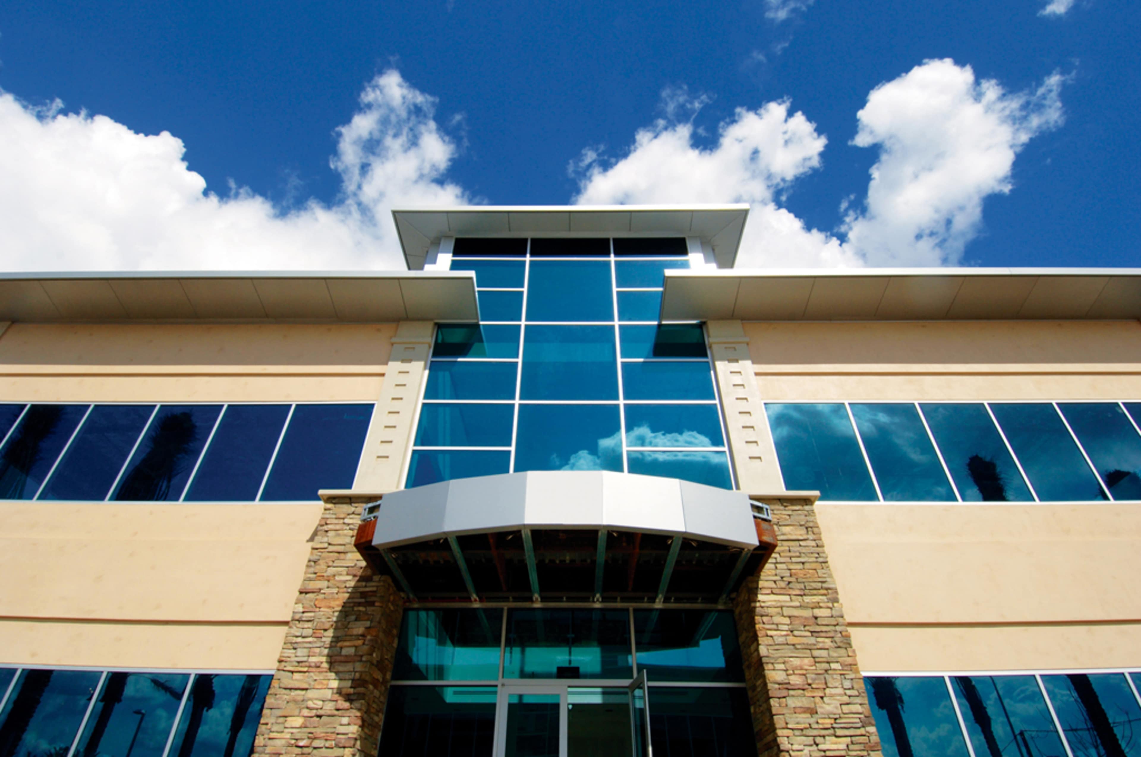 A modern building with a glass facade, featuring stone accents and a bold overhang at the entrance, set against a blue sky with clouds.