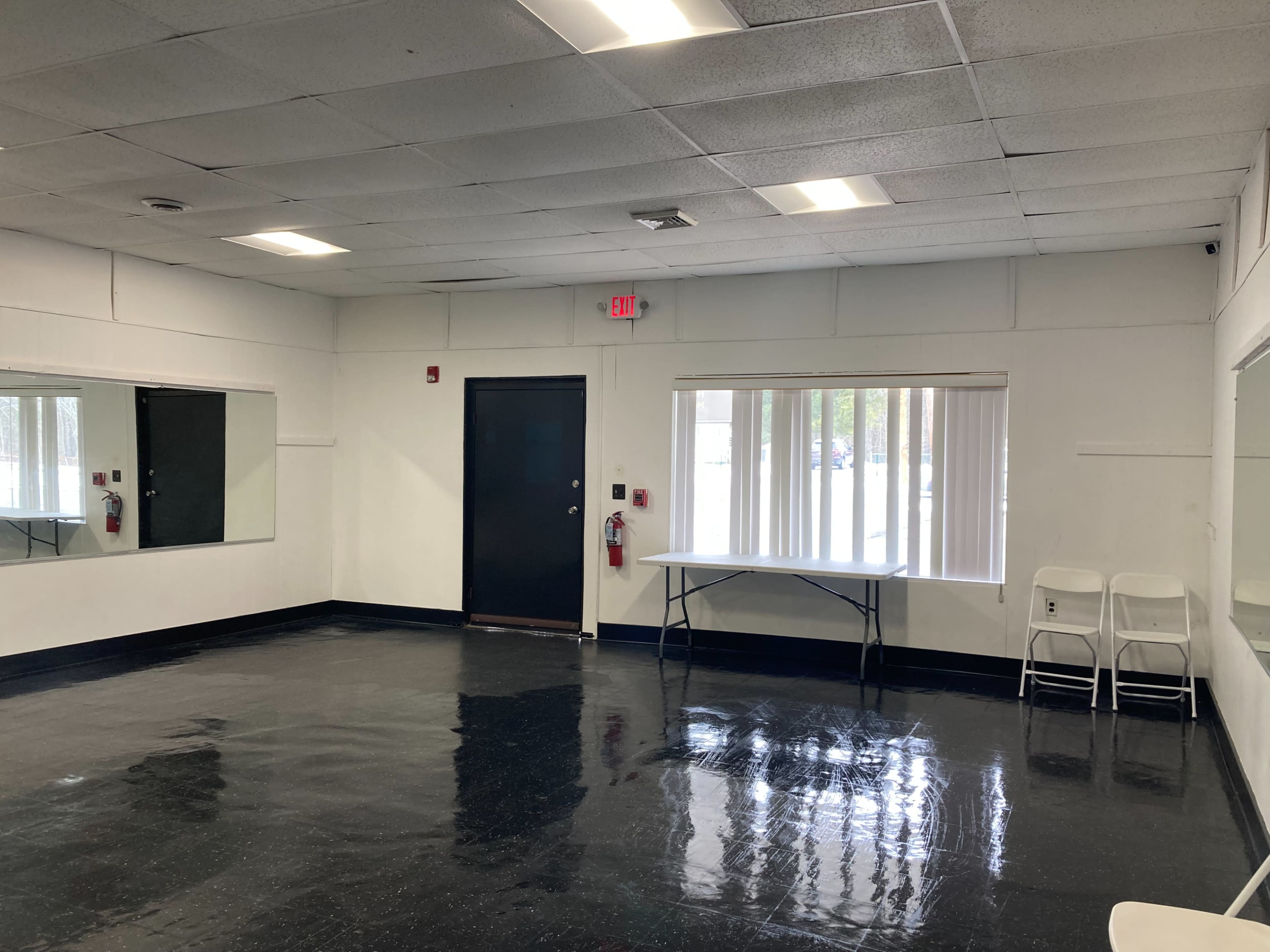 A sparsely furnished room with a mirror on one wall, a table, and two chairs, featuring glossy black flooring and white walls.