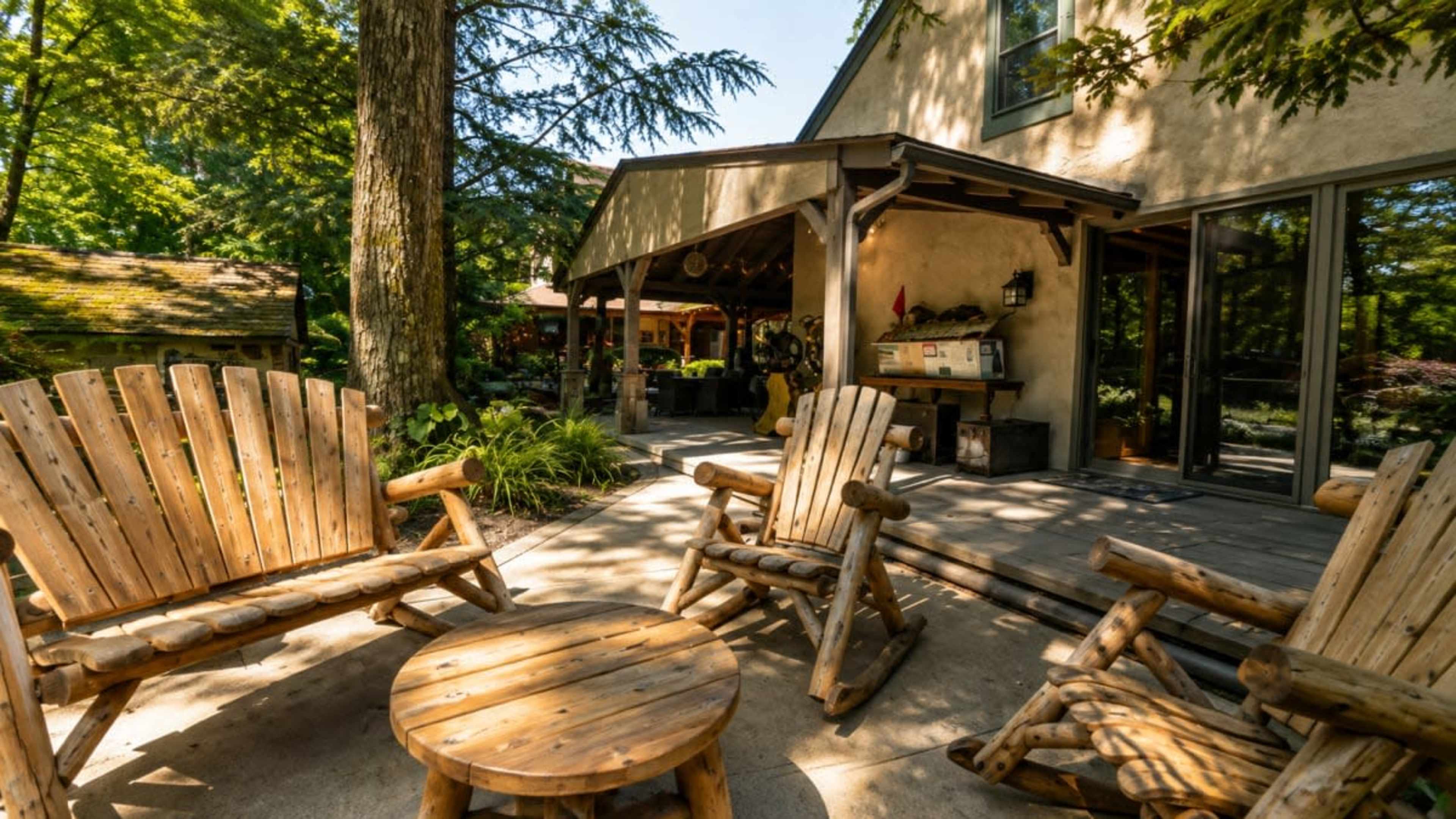 The image shows a backyard with wooden lounge chairs arranged around a small table in front of a house surrounded by trees.