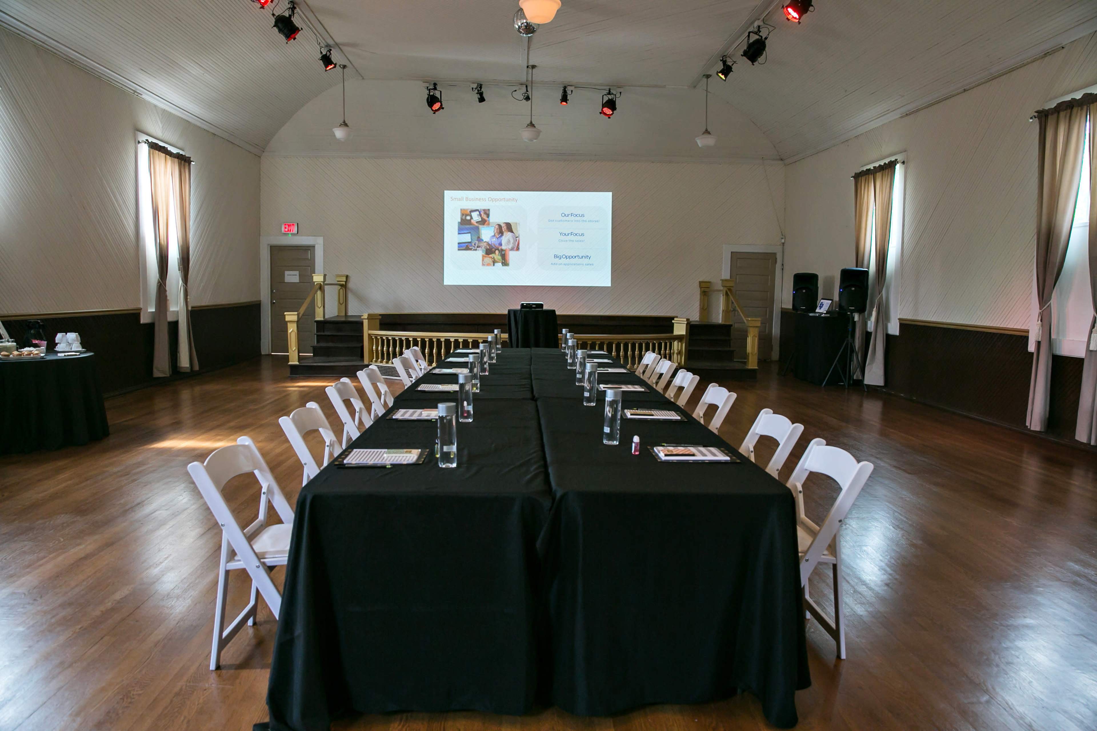 A large meeting room features a long rectangular table set with black linens and chairs, with a projector screen and audiovisual equipment at one end.