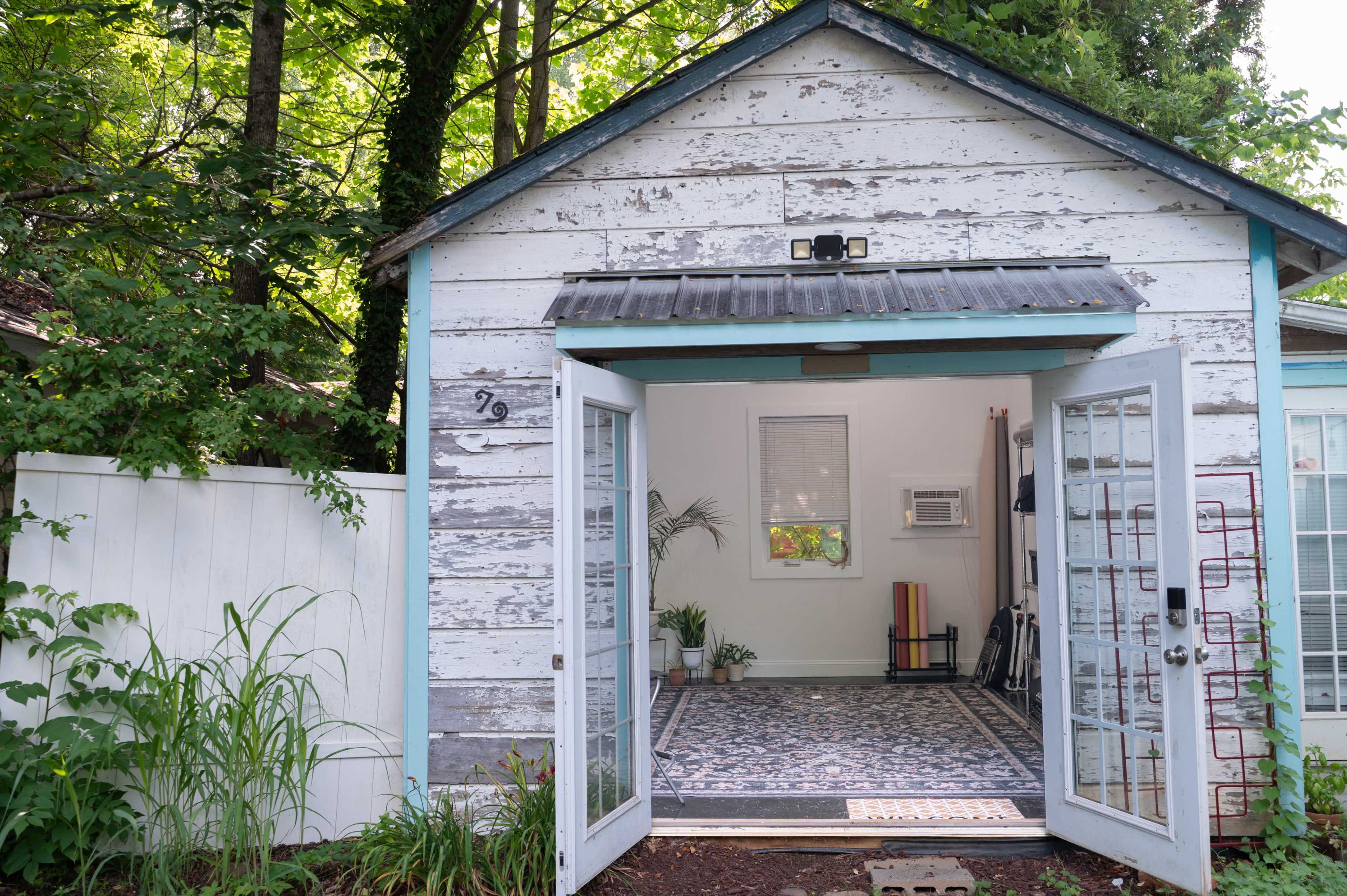 The image shows a weathered wooden shed with an open double door, revealing a neatly arranged interior featuring a floor rug and plants.