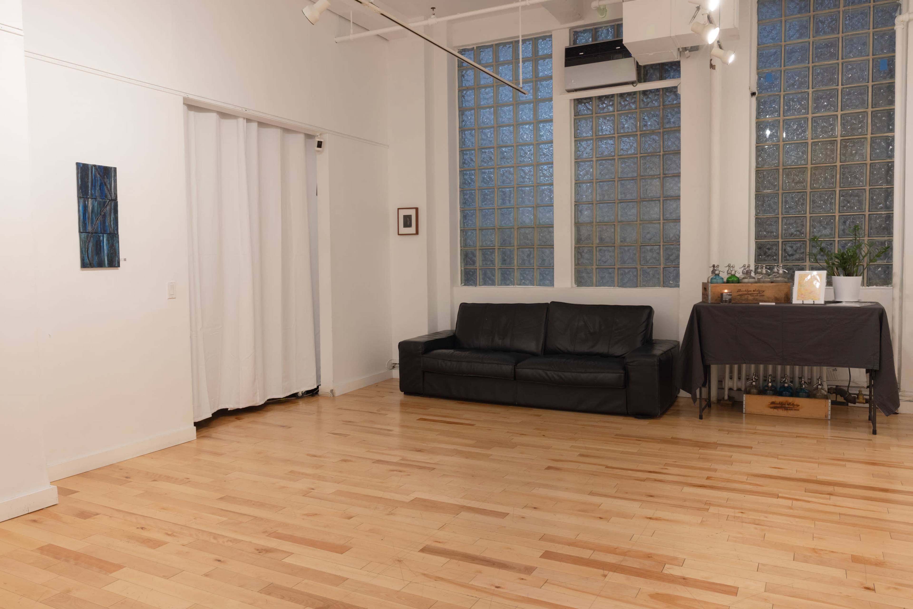 The image shows a bright, minimalist interior room featuring a black couch, a table covered with a gray cloth, a glass block wall, and wooden flooring.