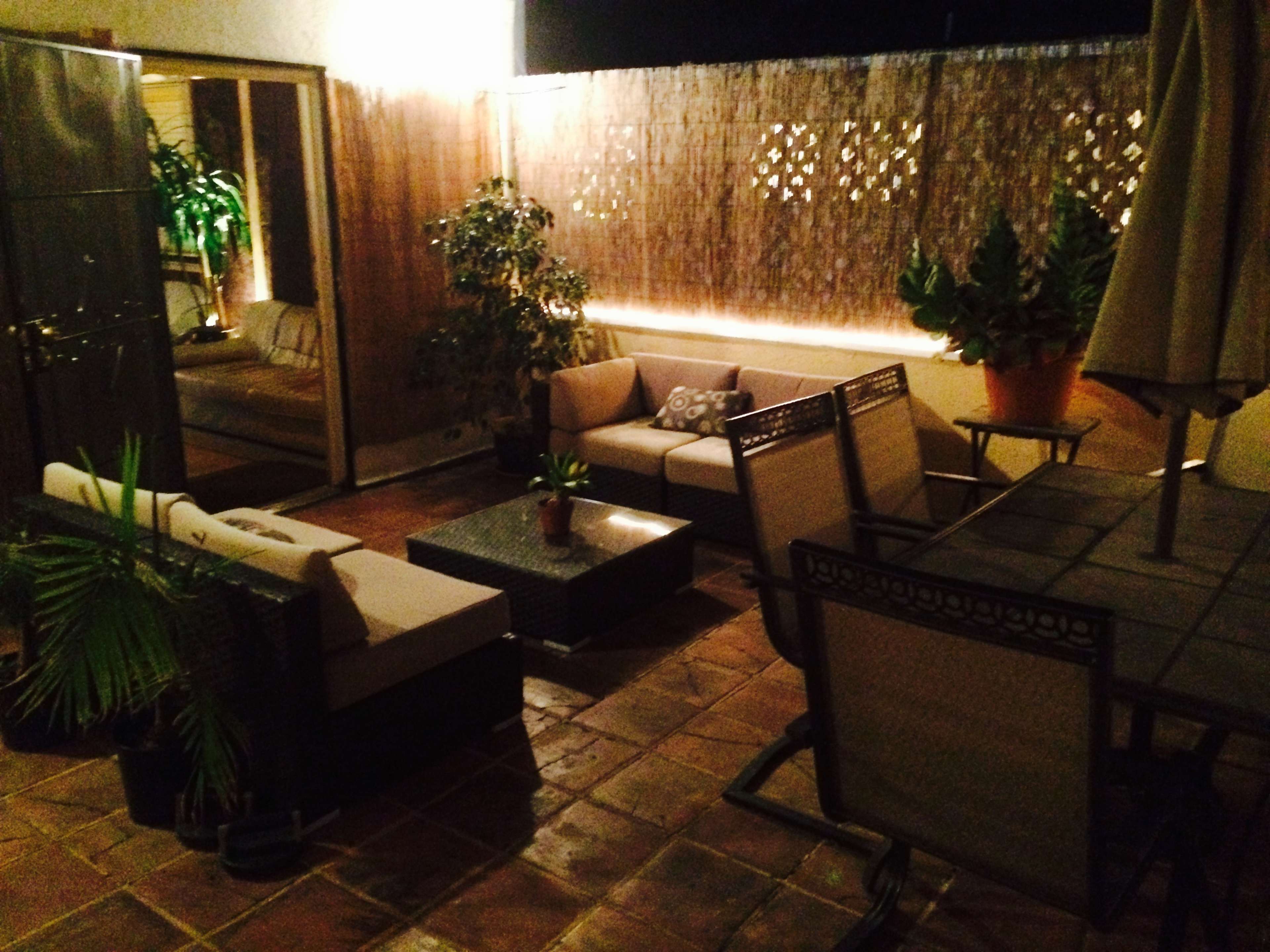 A cozy outdoor patio features a seating area with two sofas and a coffee table, surrounded by potted plants and illuminated by soft lighting.