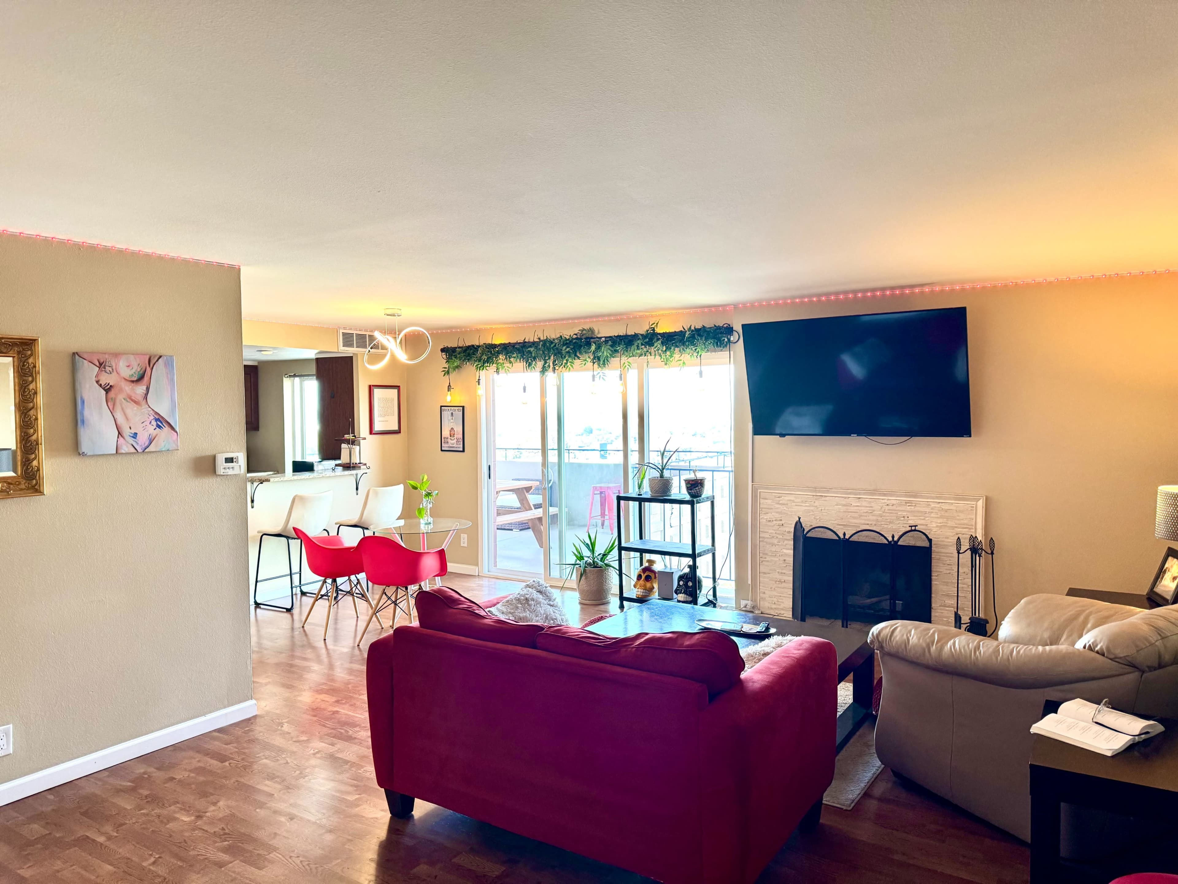 A living room features a red sofa, a beige armchair, a television mounted on the wall, and a view of a balcony through sliding glass doors.