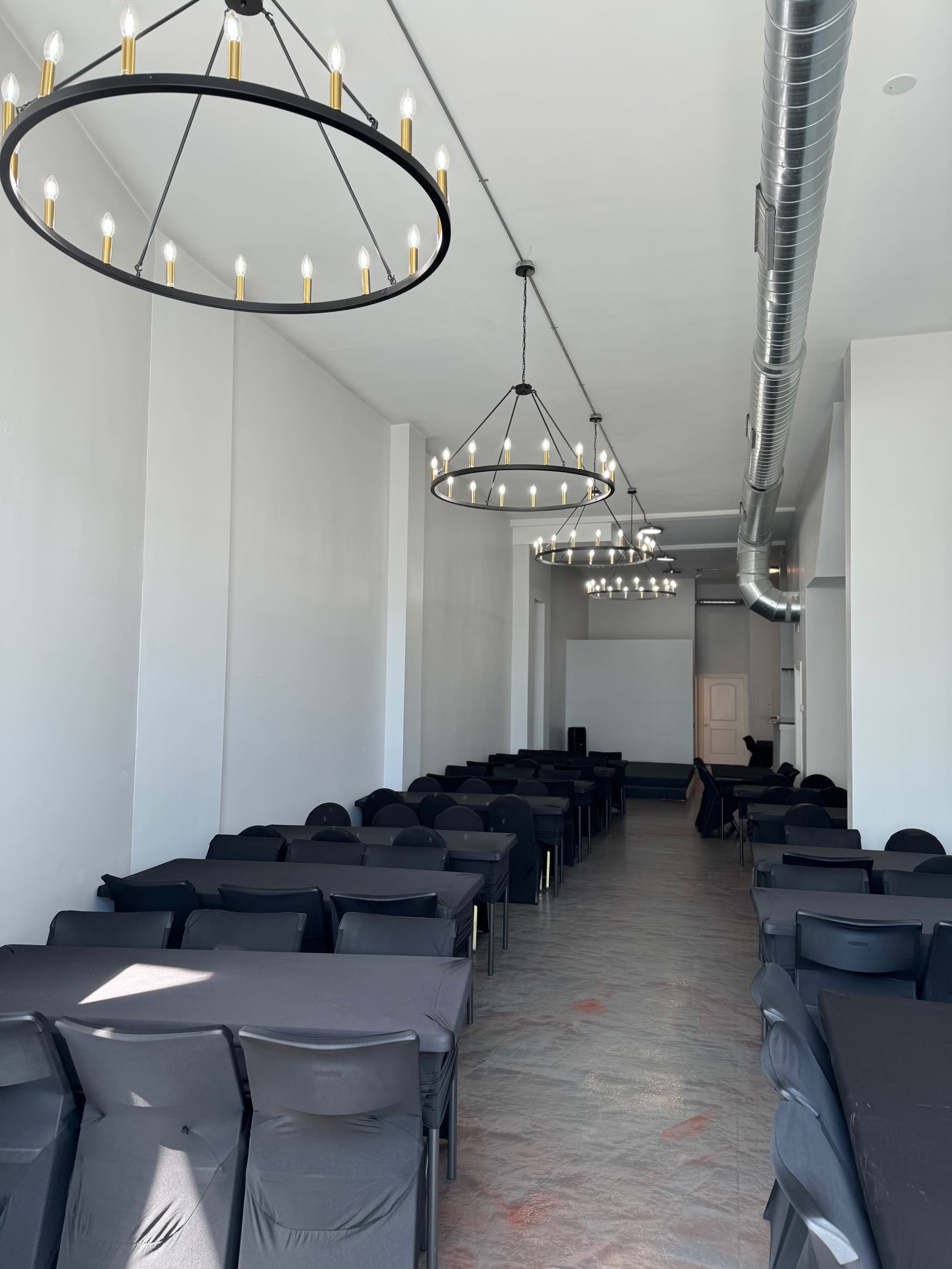 A long, narrow room with tables covered in black cloth, illuminated by round chandeliers hanging from the ceiling.